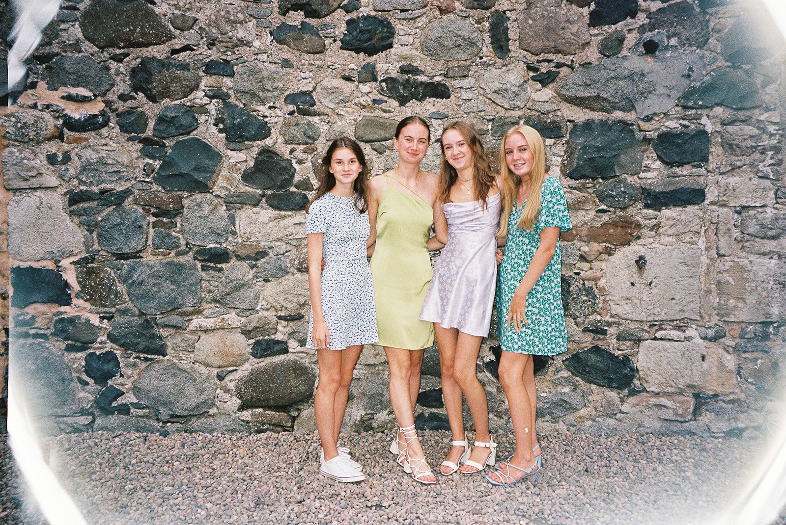Four young women standing arm-in-arm in front of a rustic stone wall, dressed in colorful summer dresses and high heels, smiling at the camera.