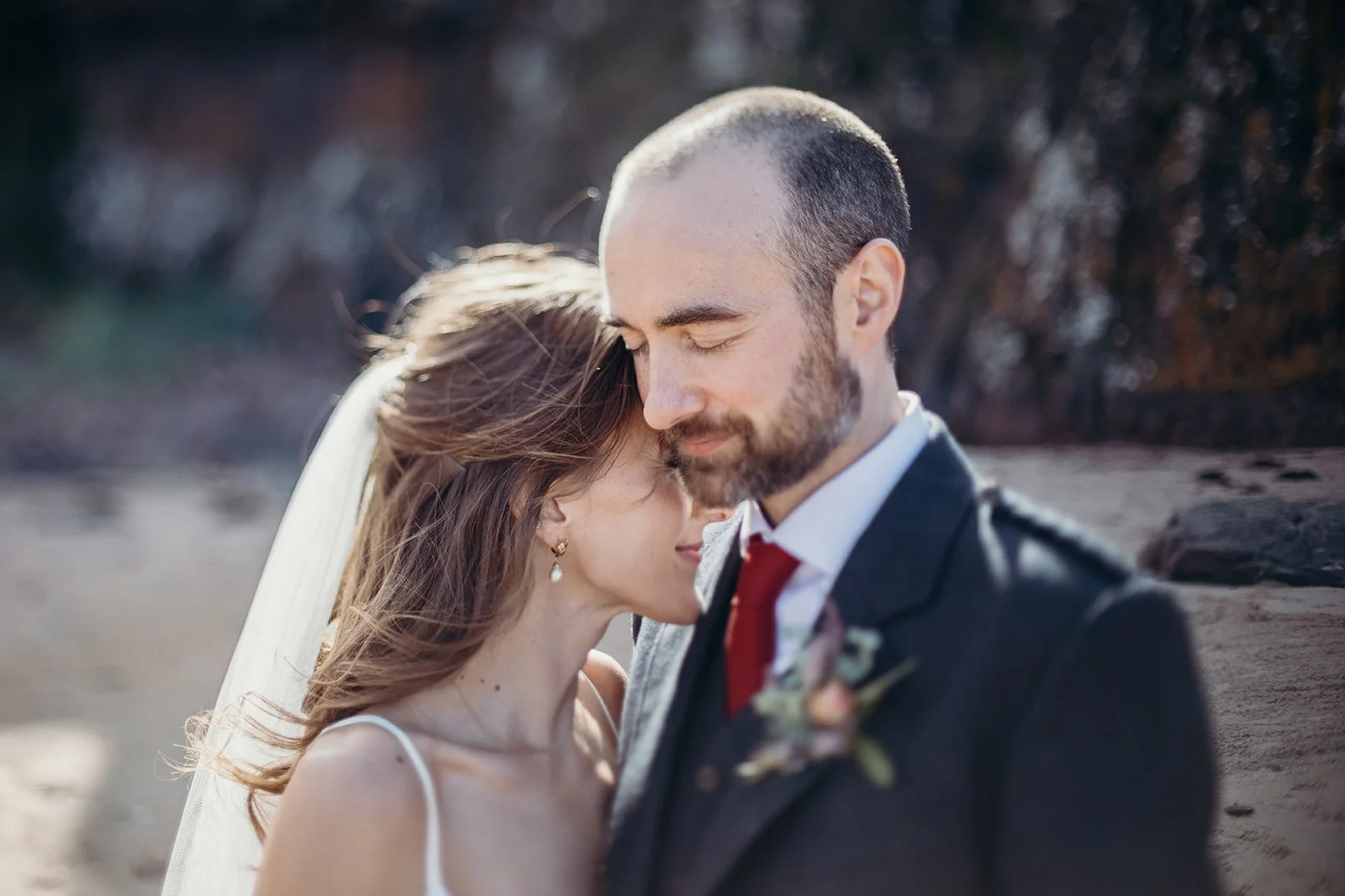 A bride and groom embracing with their foreheads touching on a beach, eyes closed, during sunset.