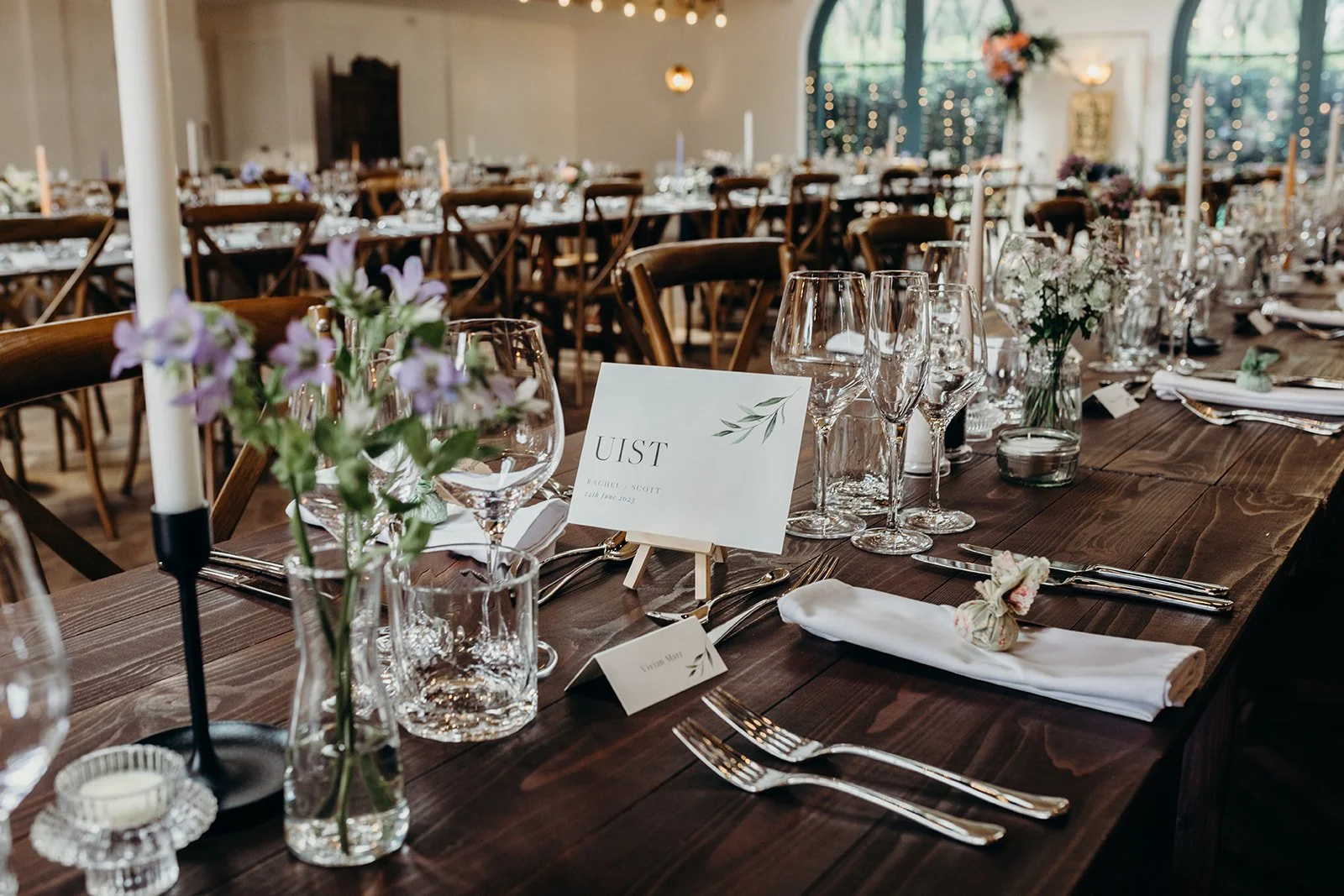 Wedding reception table decorated with flowers, glassware, and candles, with a table sign marked 'UIST' and additional place settings.