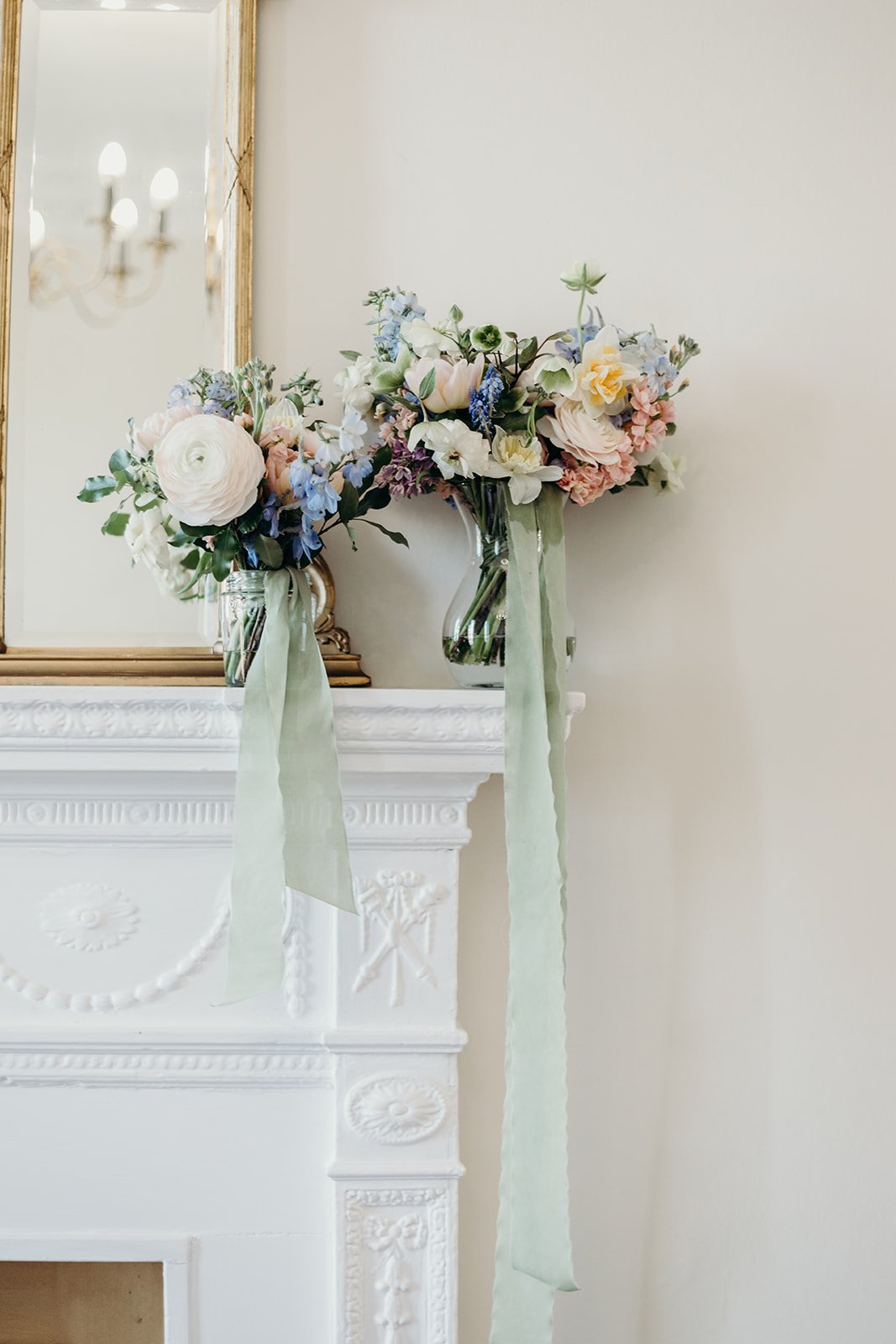 Two vases with pastel-colored flowers and long ribbons sit on a white fireplace mantel in front of a mirror with a gold frame, a chandelier reflected in the mirror behind