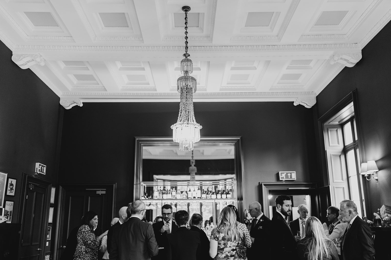 Black and white photo of a social gathering inside a grand room with high ceilings, decorative moldings, large windows, and a chandelier. People are mingling and chatting, dressed in formal attire.