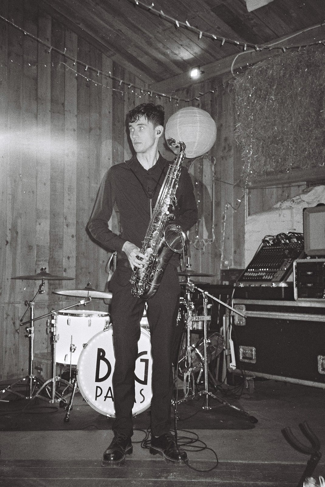 A young man holding a saxophone in a music studio with drums and sound equipment, decorated with string lights and a paper lantern.