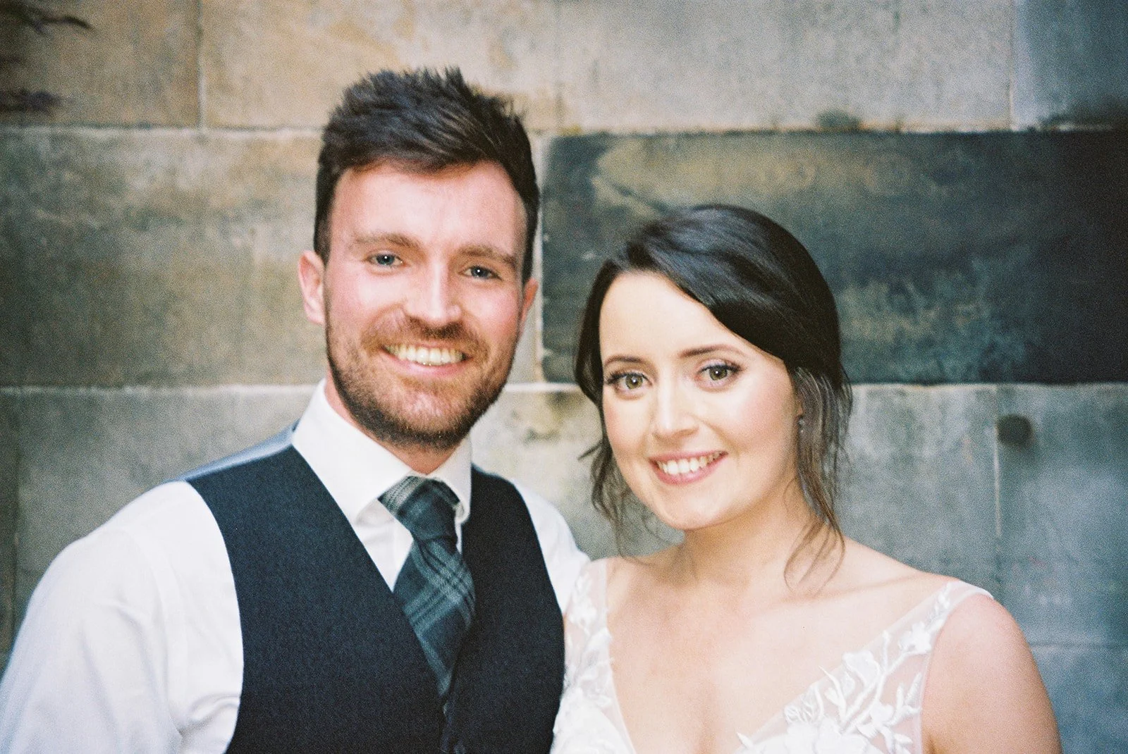 A smiling man and woman posing together indoors, with a stone wall background. The man has dark hair, a beard, and is wearing a white shirt, a dark vest, and a plaid tie. The woman has dark hair styled to the side and is wearing a white dress with la