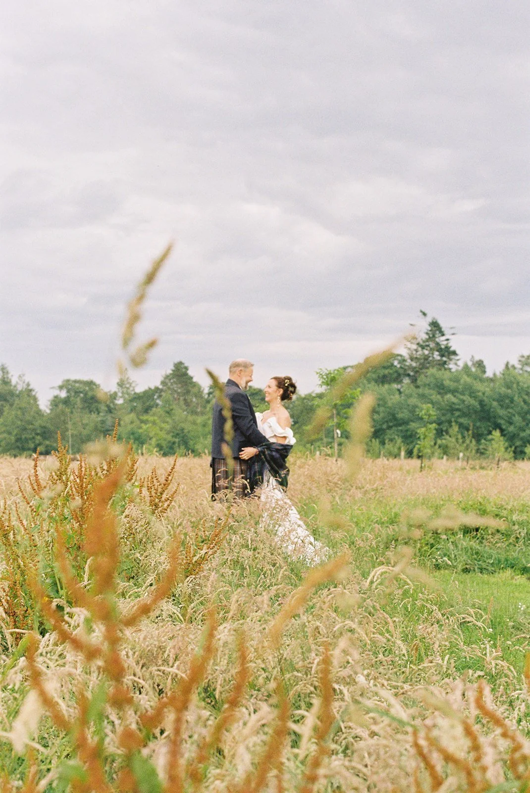 A bride and groom standing in a field of tall grass and wildflowers, holding hands and looking at each other, with trees and cloudy sky in the background.