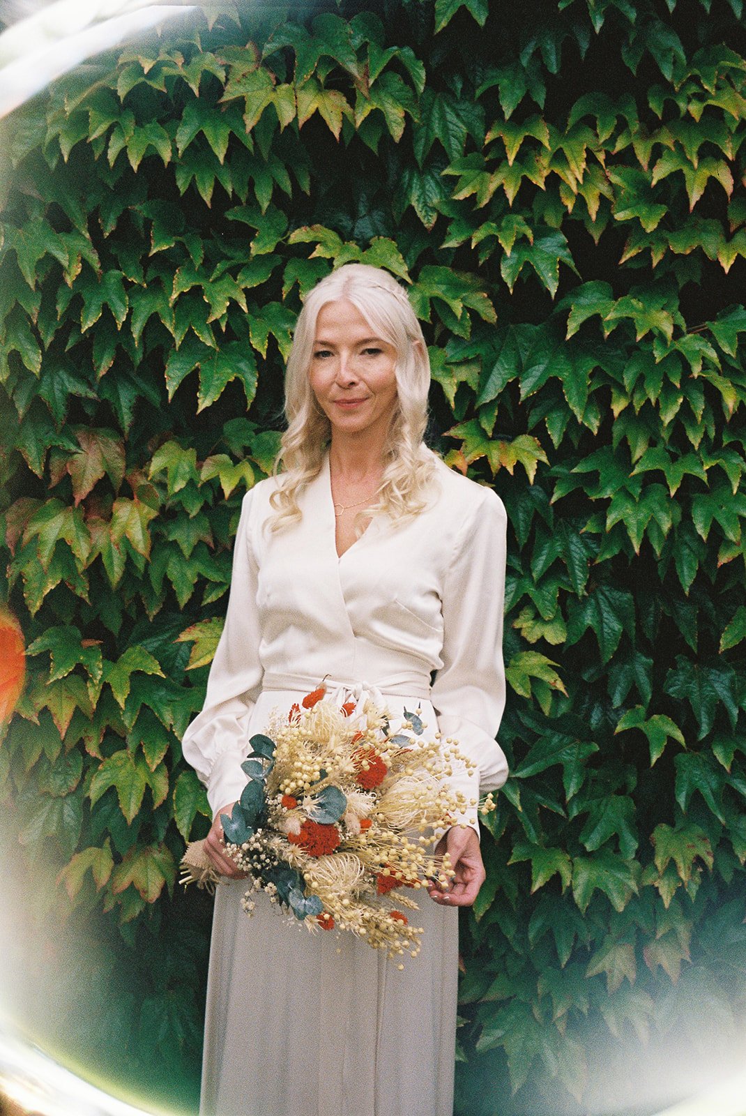 A woman with long blond hair wearing a cream-colored blouse and gray skirt holding a bouquet of dried flowers with green leaves, standing in front of lush green ivy covering a wall.