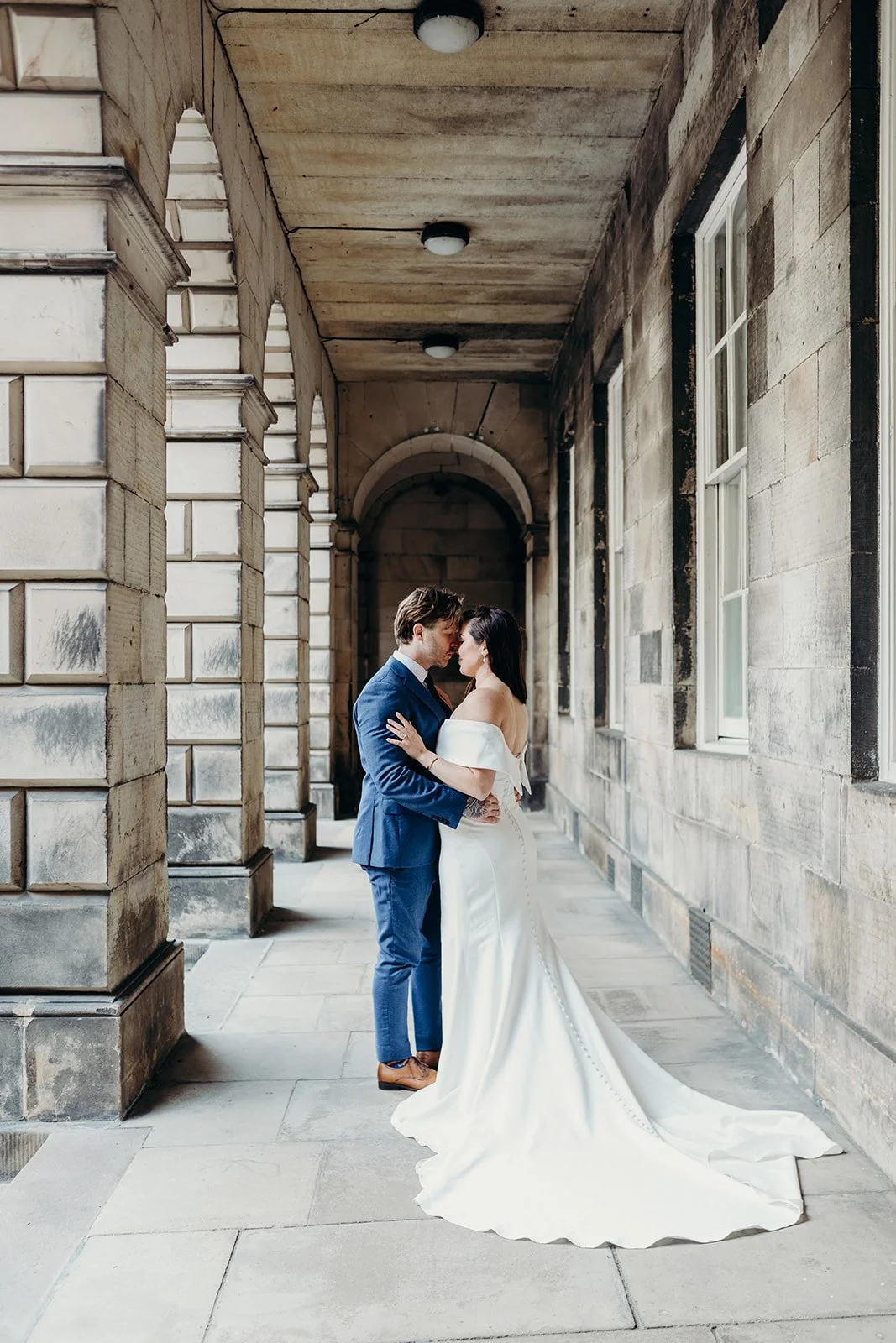 Bride and groom standing closely together on a stone walkway under a covered archway, with the bride in a white wedding dress and the groom in a blue suit, facing each other with their foreheads touching.
