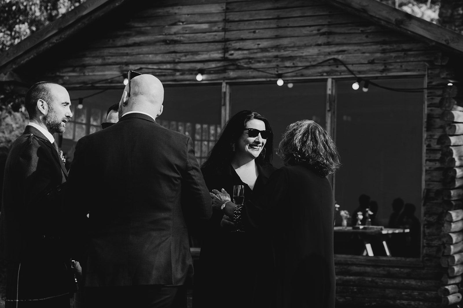 A woman with dark hair and sunglasses smiling while speaking to an older woman, with three men in suits nearby, in an outdoor wooden shelter at a social gathering or celebration.