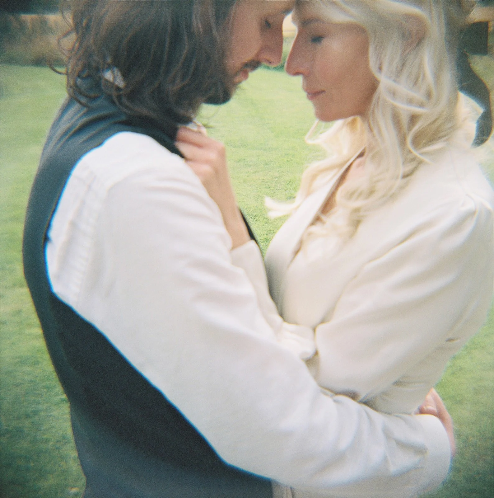 A man and woman in an intimate embrace with their foreheads touching outdoors in a grassy area.