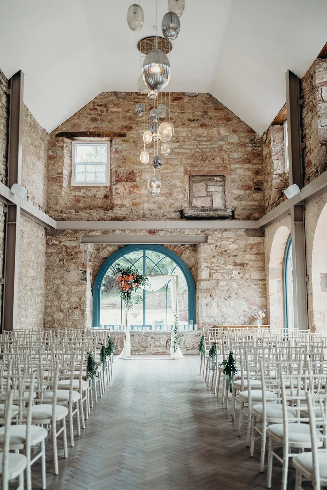 Indoor wedding venue with white chairs decorated with greenery, a floral arch, large arched window, brick walls, wooden floor, hanging glass pendant lights, and small windows in a high vaulted ceiling.