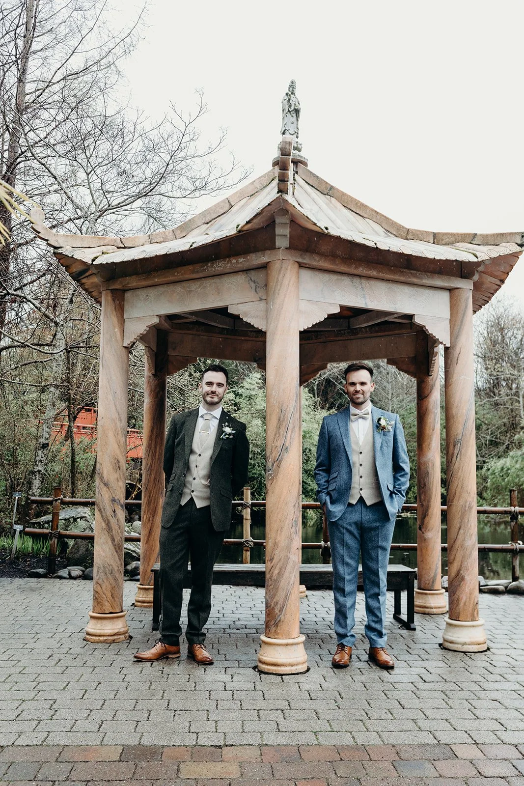 Two men in formal suits standing under a wooden pagoda by a pond, with trees in the background.