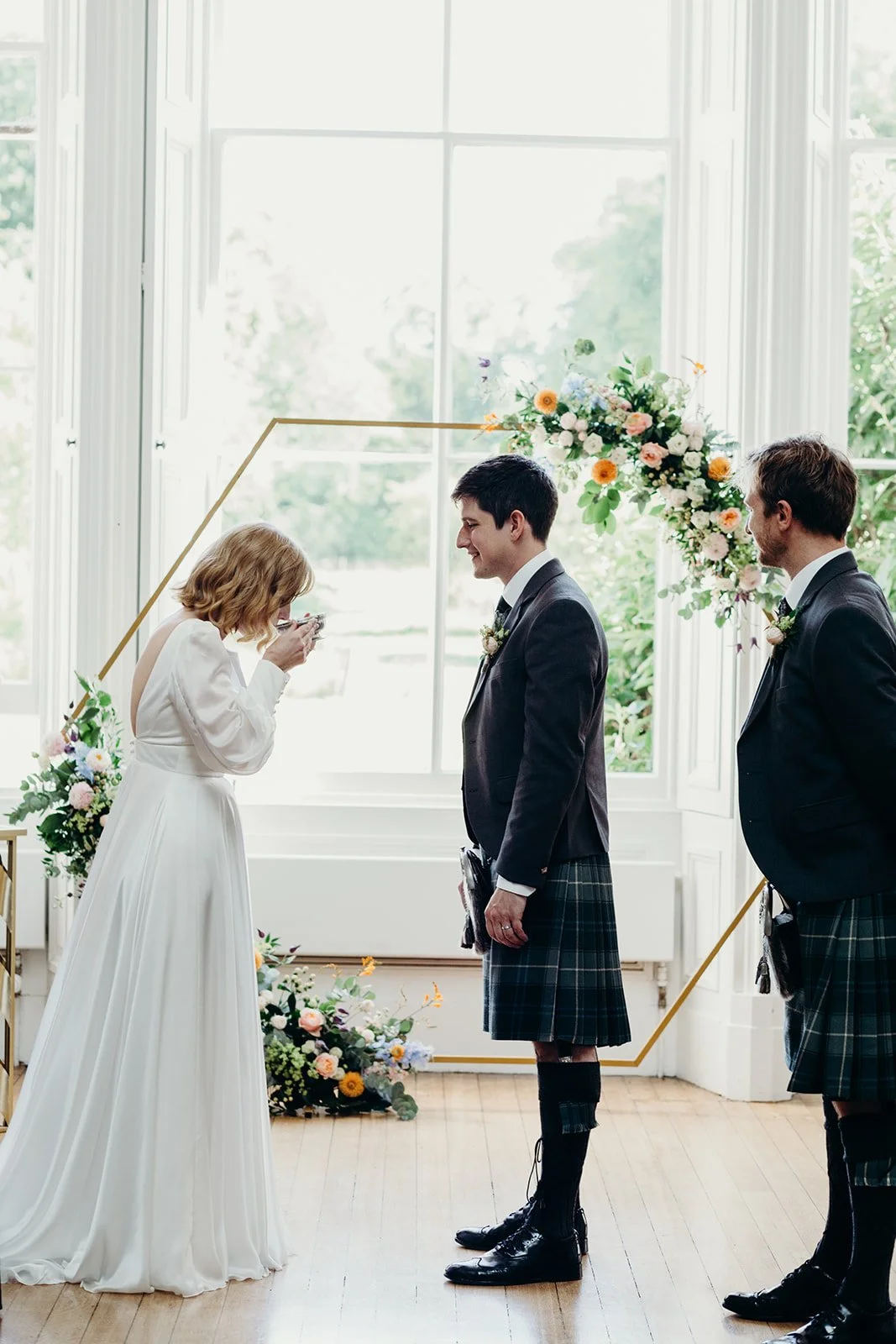 A wedding ceremony showing a bride, groom, and another man in a room with large windows and floral decorations.