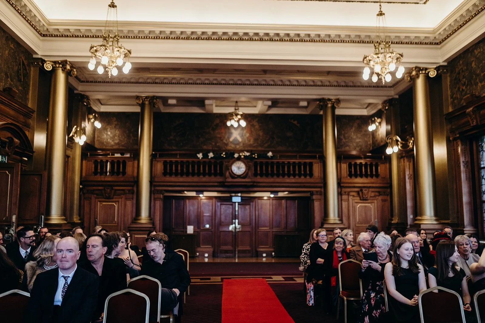 People sitting in chairs inside a grand, wood-paneled room with high ceilings, large gold columns, chandeliers, and a red carpet ran down the center.