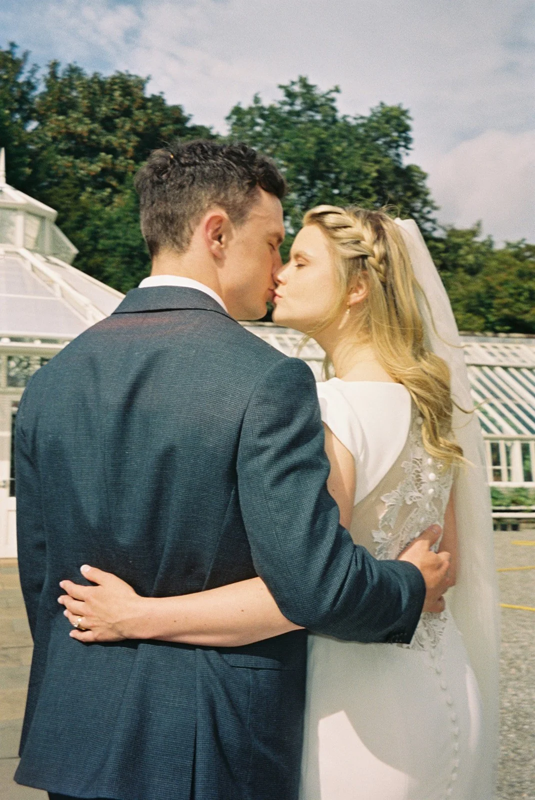 A bride and groom are sharing a kiss outdoors on their wedding day, with the bride wearing a white dress with lace details and the groom in a dark suit. They are embracing each other, with trees and a gazebo visible in the background.