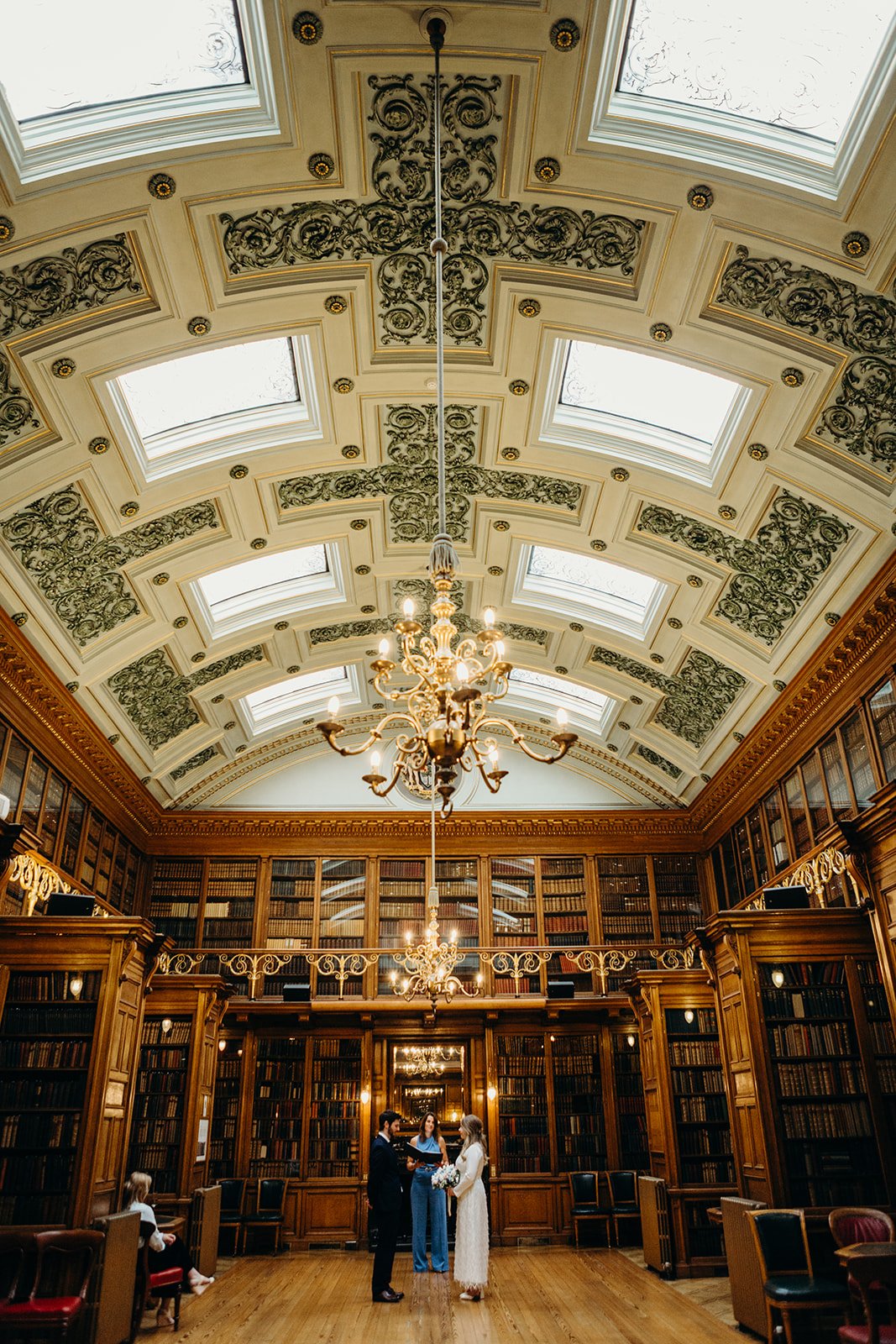 A wedding ceremony taking place in a grand, historic library with ornate ceiling, chandeliers, wood-paneled walls, and tall bookshelves. The bride and groom are facing each other with an officiant between them, while a person sits on a chair in the foreground.