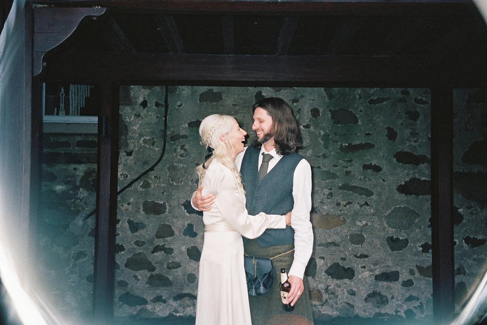 A couple, a bride with long blond hair and a groom with dark, shoulder-length hair, sharing a joyful moment and smiling at each other while dancing at their wedding reception in front of a stone wall. The groom is holding a beer bottle, and the bride