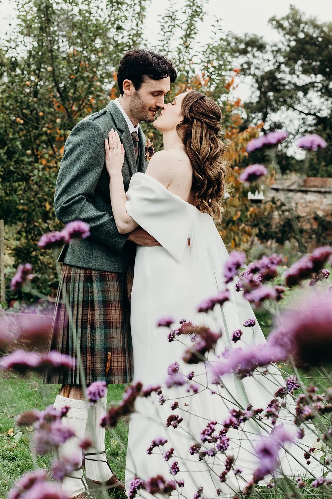 A newlywed couple embracing outdoors, surrounded by pink and purple flowers, with trees and a rustic building in the background.