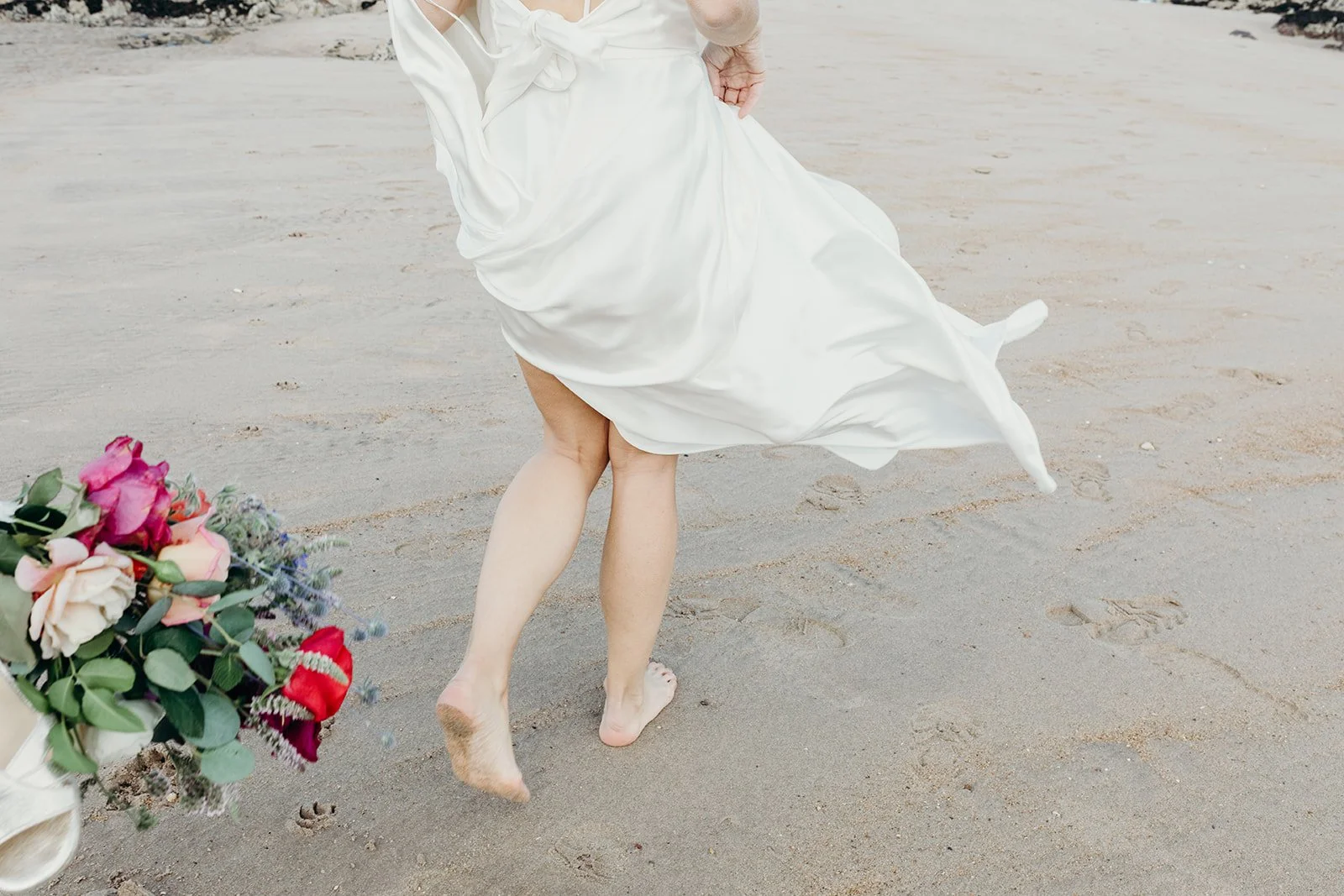 A woman in a white dress walking barefoot on a sandy beach, holding a bouquet of flowers.