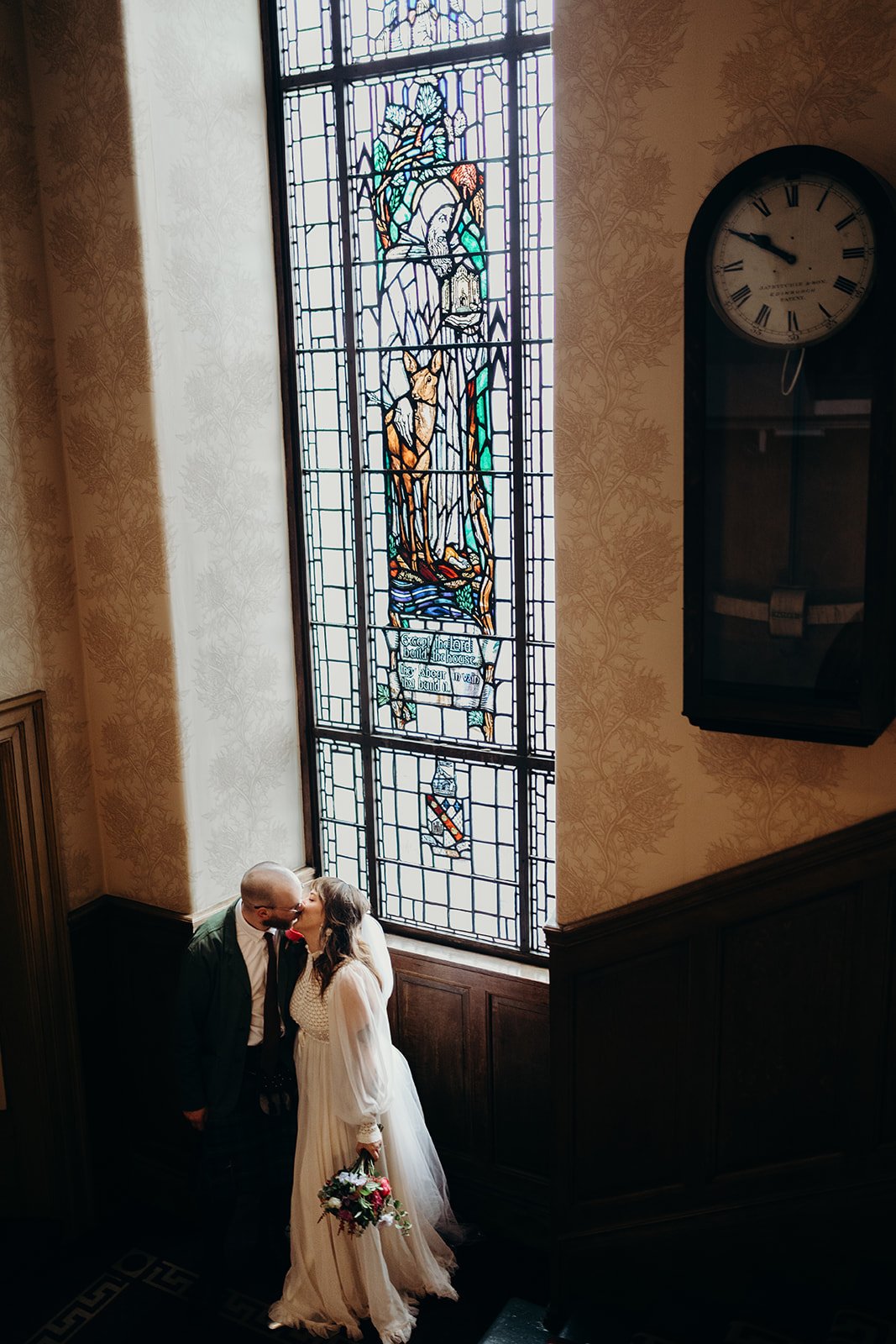A wedding couple kissing near a stained glass window in a church, with a clock on the wall nearby.