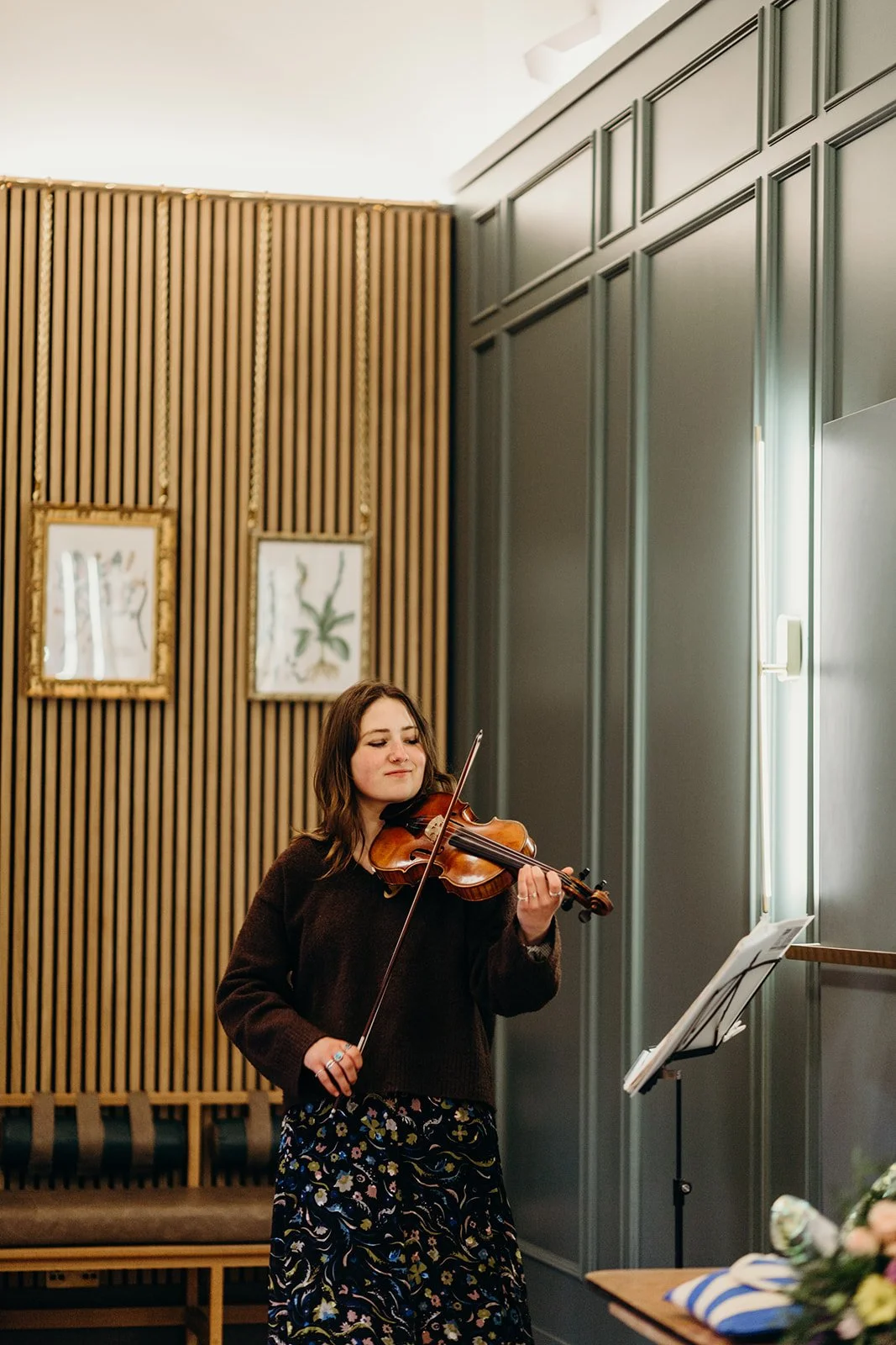 A woman playing the violin in a room with contemporary and traditional decor, including a wood-paneled wall and framed botanical artwork.