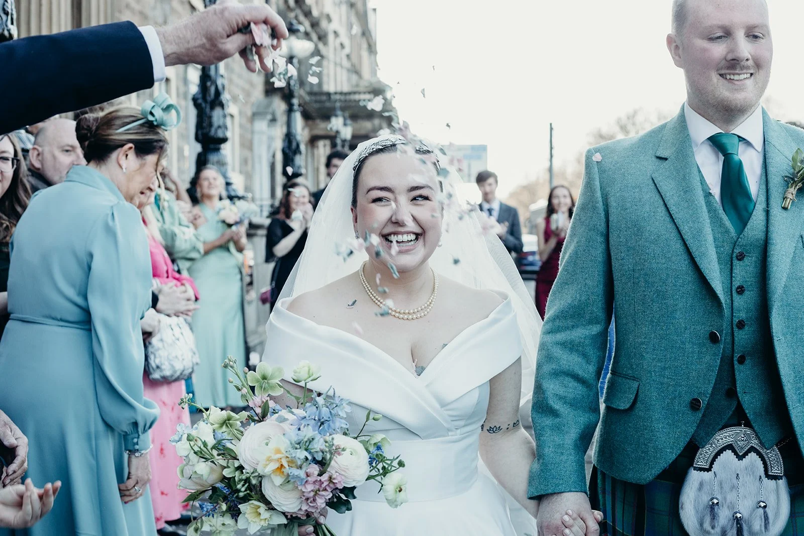 A bride and groom smiling as they walk hand-in-hand through a crowd during their wedding celebration, with confetti falling around them.