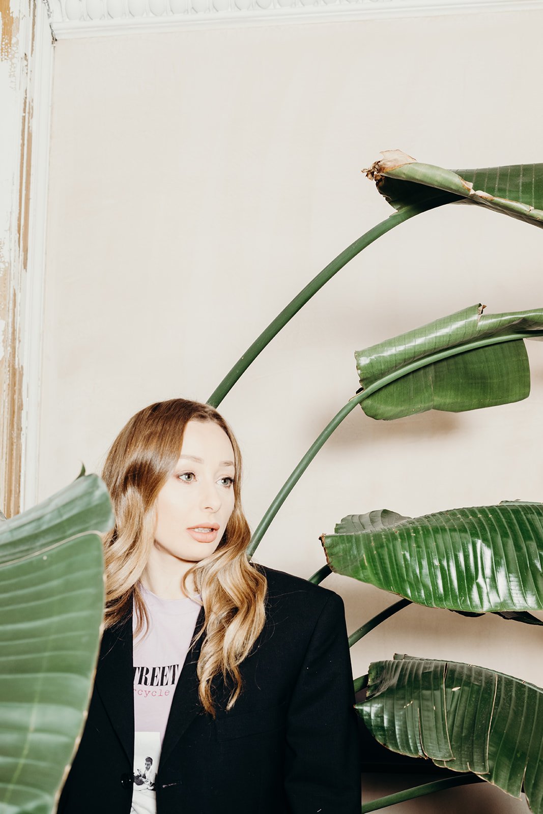 A woman with wavy blond hair, wearing a black blazer, is partially obscured by large green tropical leaves in an indoor setting.