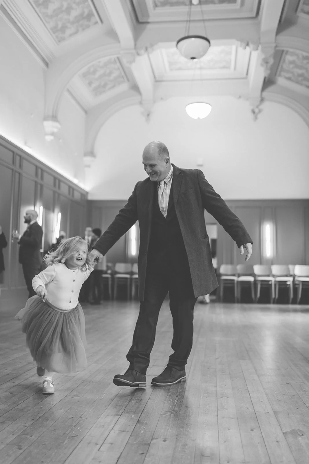 A man and a young girl dancing and smiling in a large, elegant room with high ceilings and ornate details.