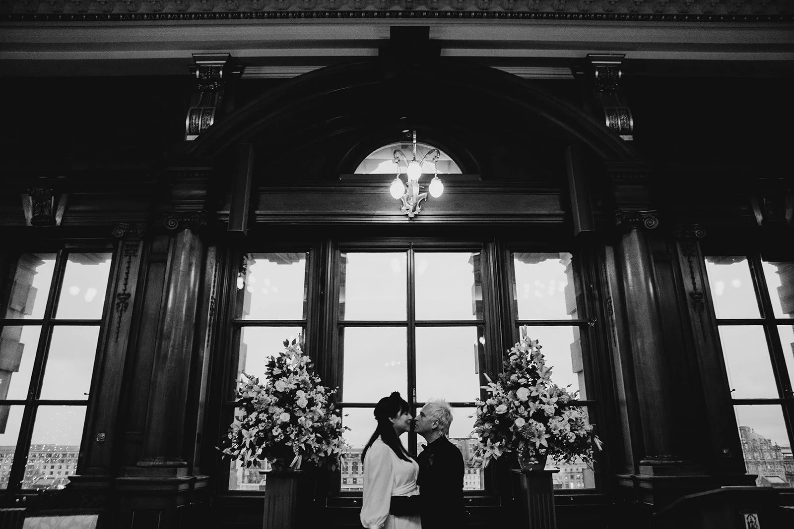Two women are standing close and kissing in front of large windows with cityscape view, decorated with floral arrangements.