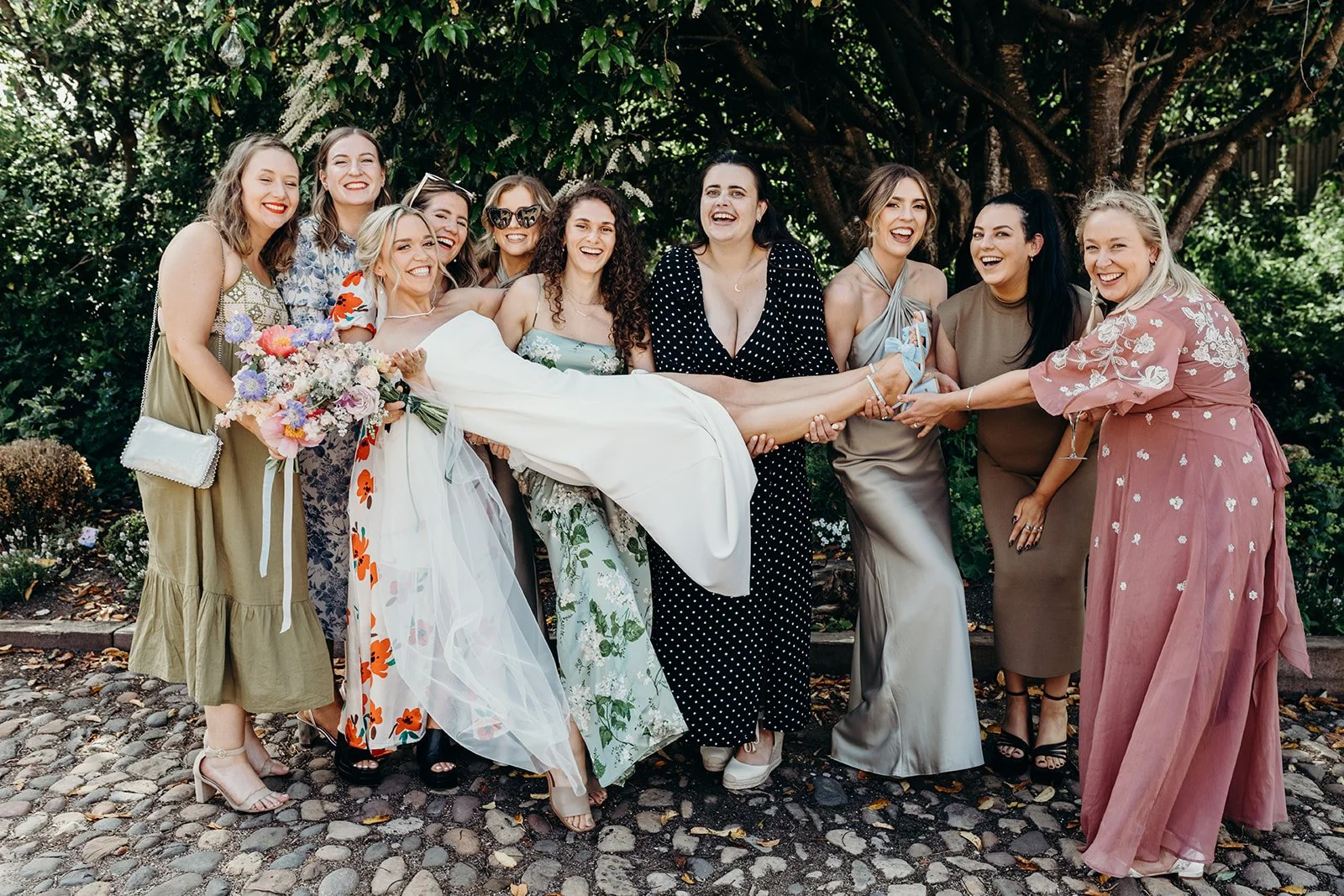 Group of women celebrating a wedding outdoors, with one woman in a wedding dress being carried by friends, all smiling and laughing.