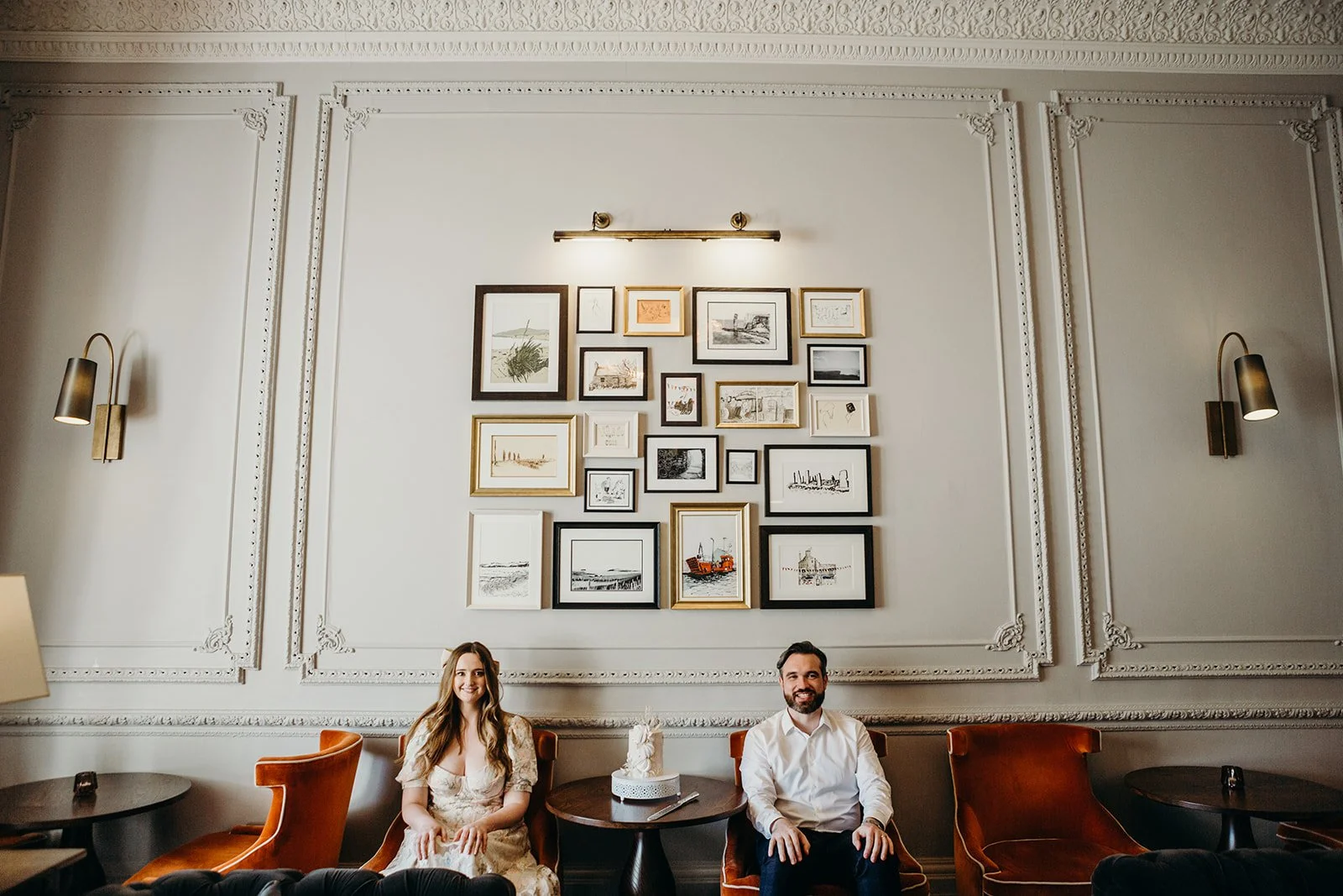 A man and woman sitting on orange chairs in a stylish room with ornate white walls, surrounded by framed pictures and artwork, with a small table holding a cake in front of them.
