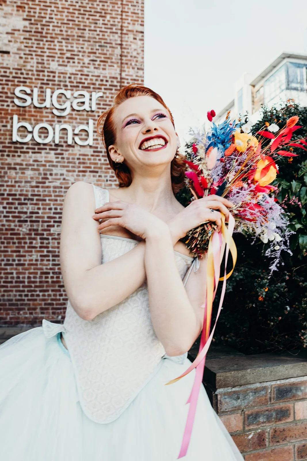 A woman in a white wedding dress holding a colorful bouquet of flowers, smiling joyfully outside near a brick wall with a sign that reads 'sugar bonda'.