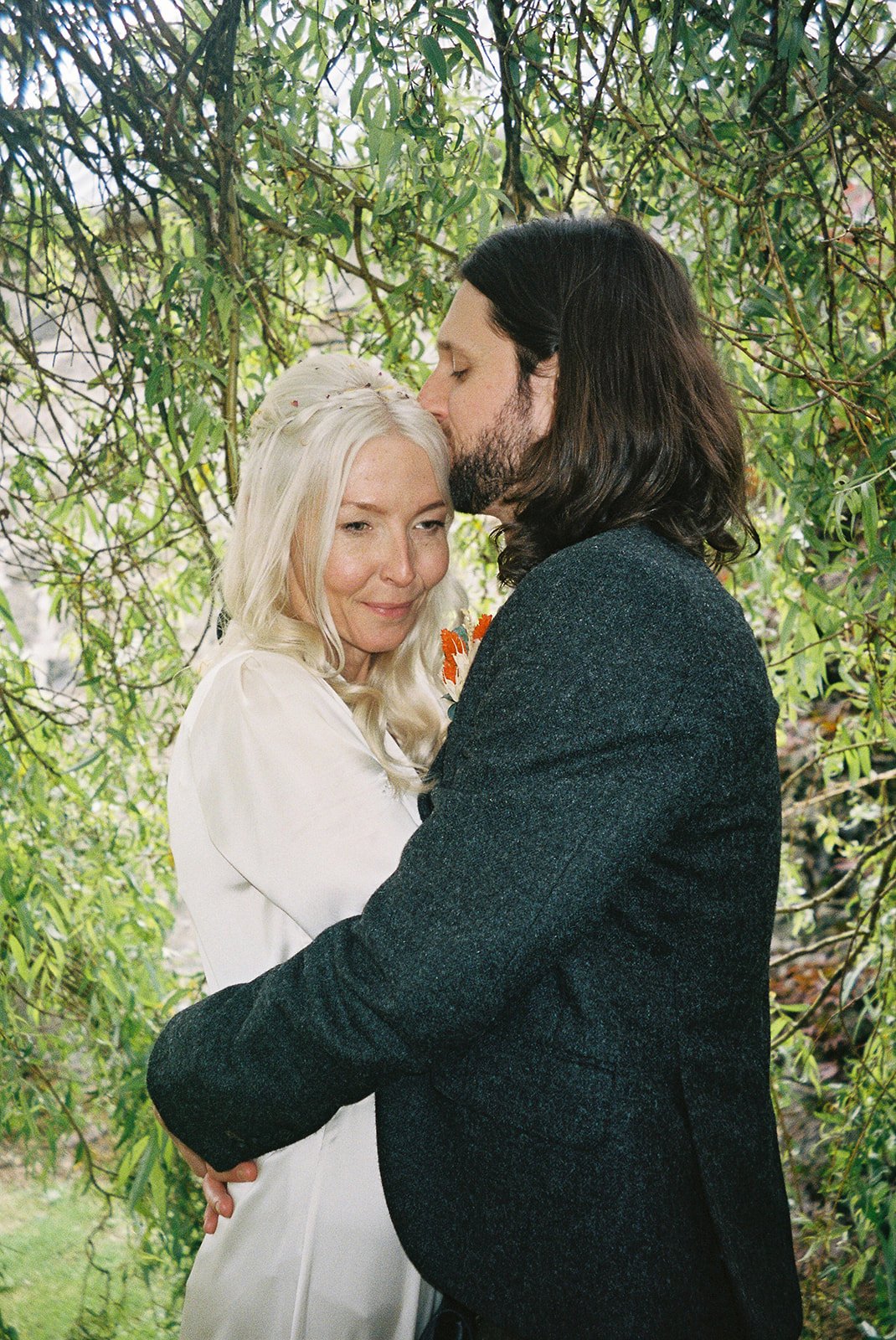A couple dressed in wedding attire standing close together and sharing a tender moment in a lush green outdoor setting. The man is kissing the woman's forehead while she smiles softly with her eyes closed.
