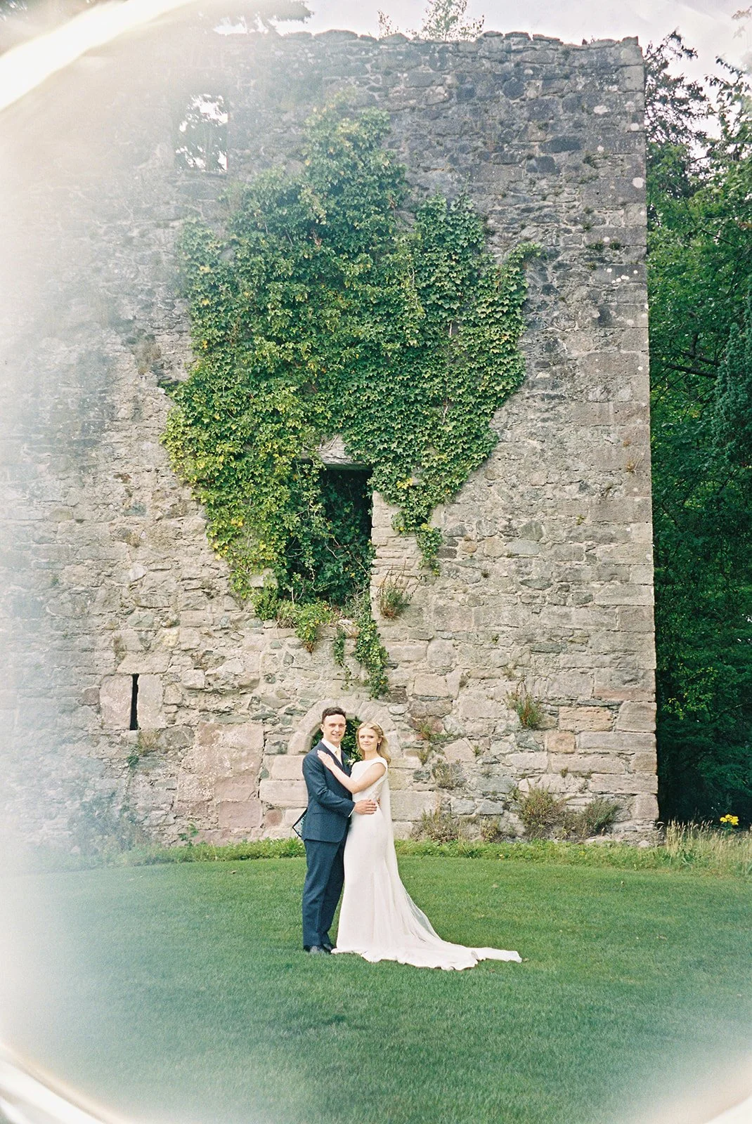 A bride and groom in wedding attire standing together on grass in front of an old stone wall with ivy growing on it.