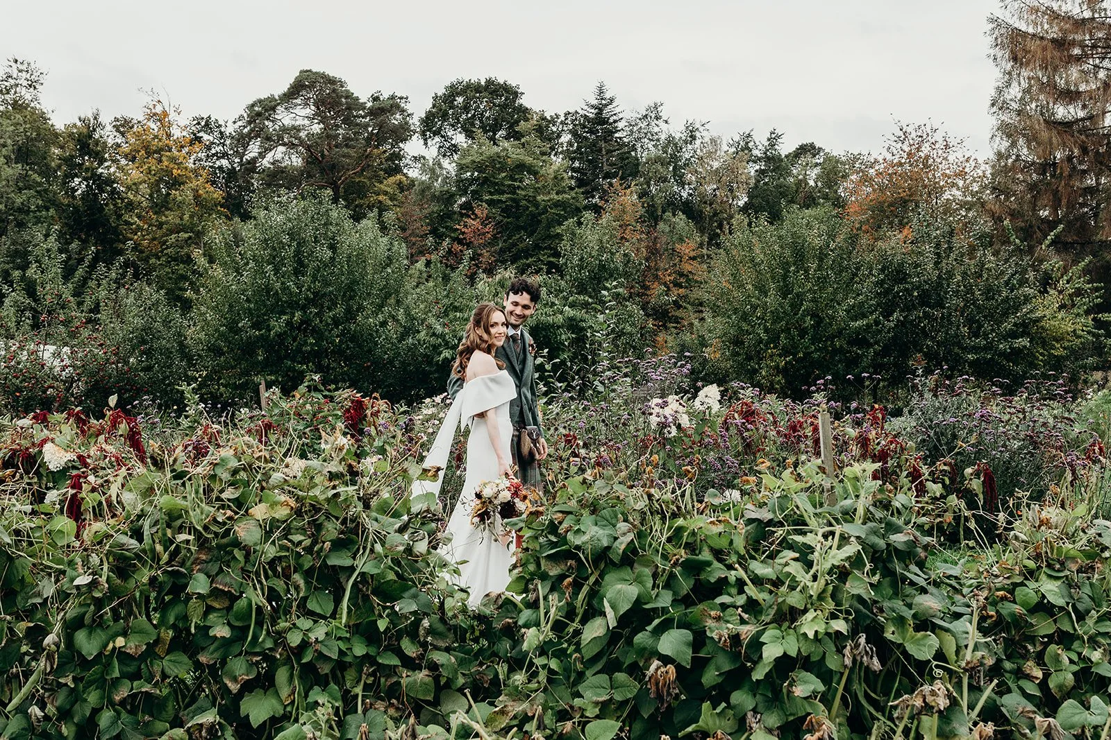 A couple in wedding attire standing together in a lush garden with dense green trees and colorful autumn foliage in the background.