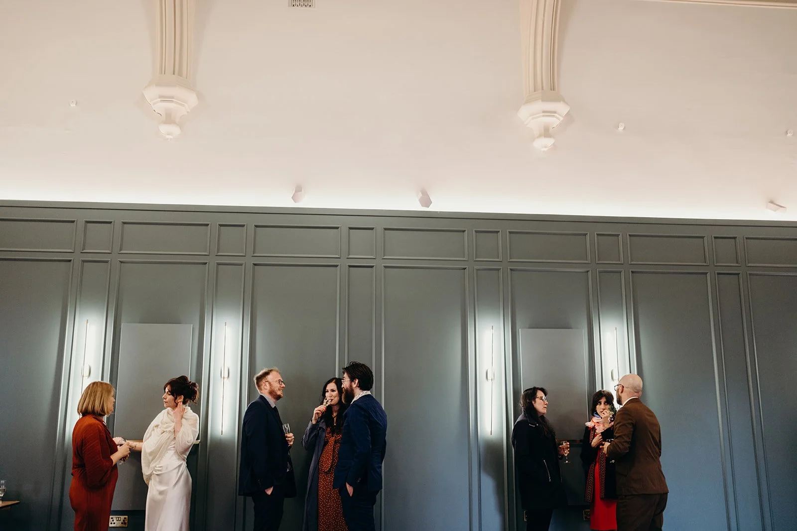 People socializing and talking at a formal indoor event in a room with gray paneled walls and decorative ceiling features.