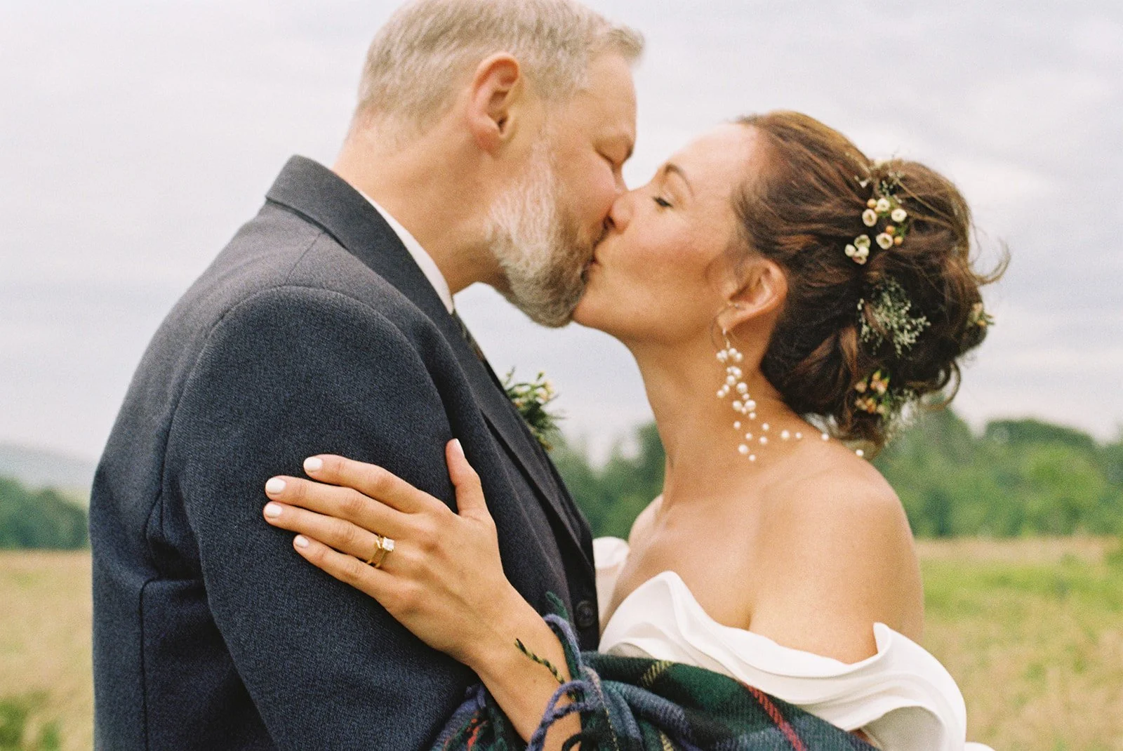 A couple kissing outdoors, celebrating their wedding, with the woman wearing a white off-the-shoulder dress and the man in a dark suit, in a field with trees in the background.