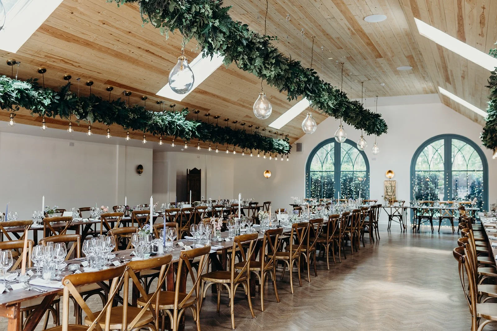 Wedding reception setup in a decorated hall with wooden chairs and tables set with glassware and cutlery, large arched windows, hanging string lights, and greenery decorations on the ceiling.