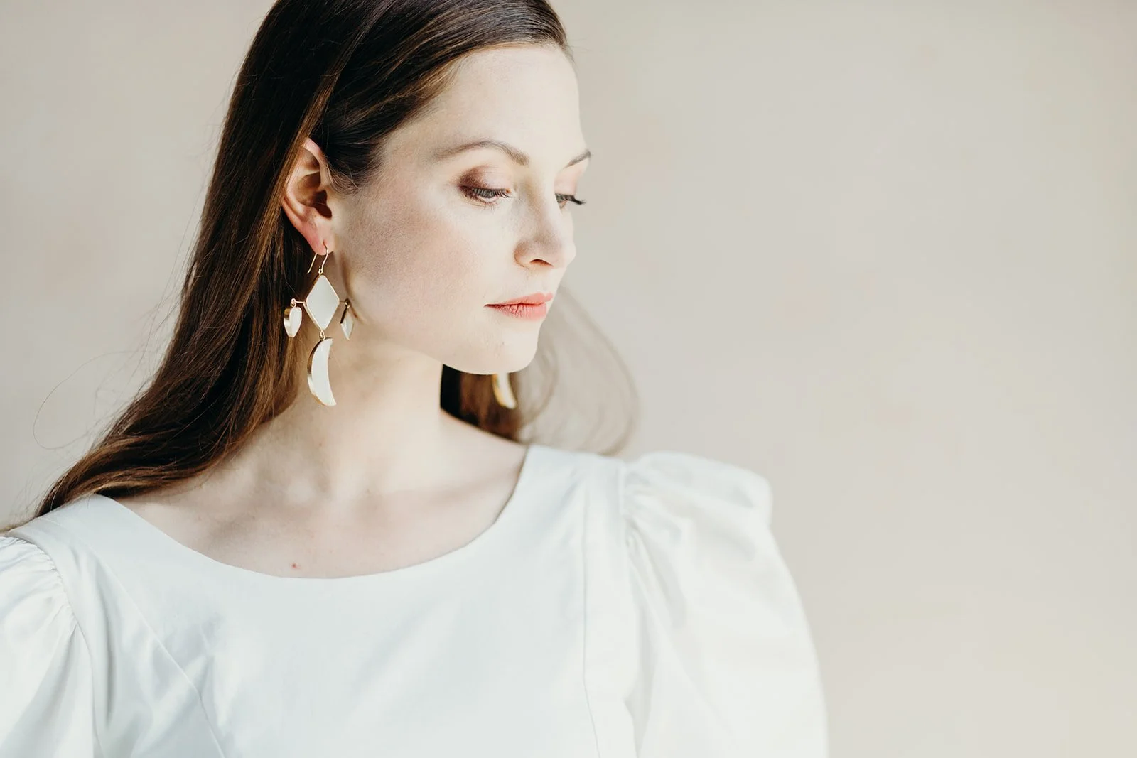 A woman with long brown hair wearing large, gold and white earrings and a white top, looking to the left with a neutral expression against a plain, light-colored background.
