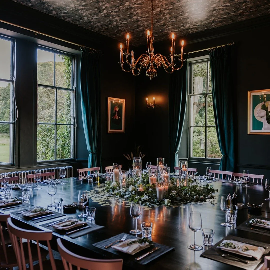 Elegant dining room with dark walls, large windows with blue curtains, a chandelier and wall sconces, a long table with place settings, glasses, napkins, and a floral centerpiece with candles.