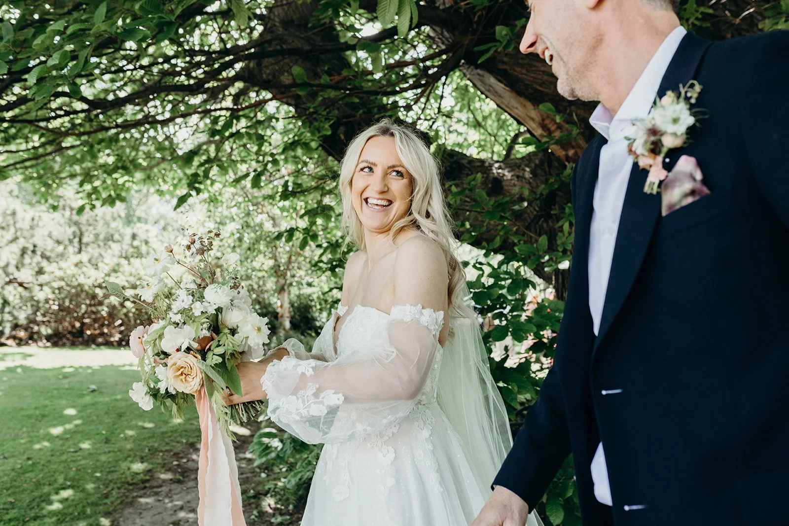A bride holding a bouquet smiling at a man, outside under a tree on a sunny day.