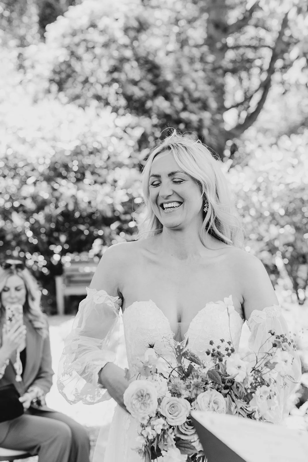 Black and white photo of a bride smiling while holding a bouquet of flowers outdoors, with a guest taking a photo in the background.