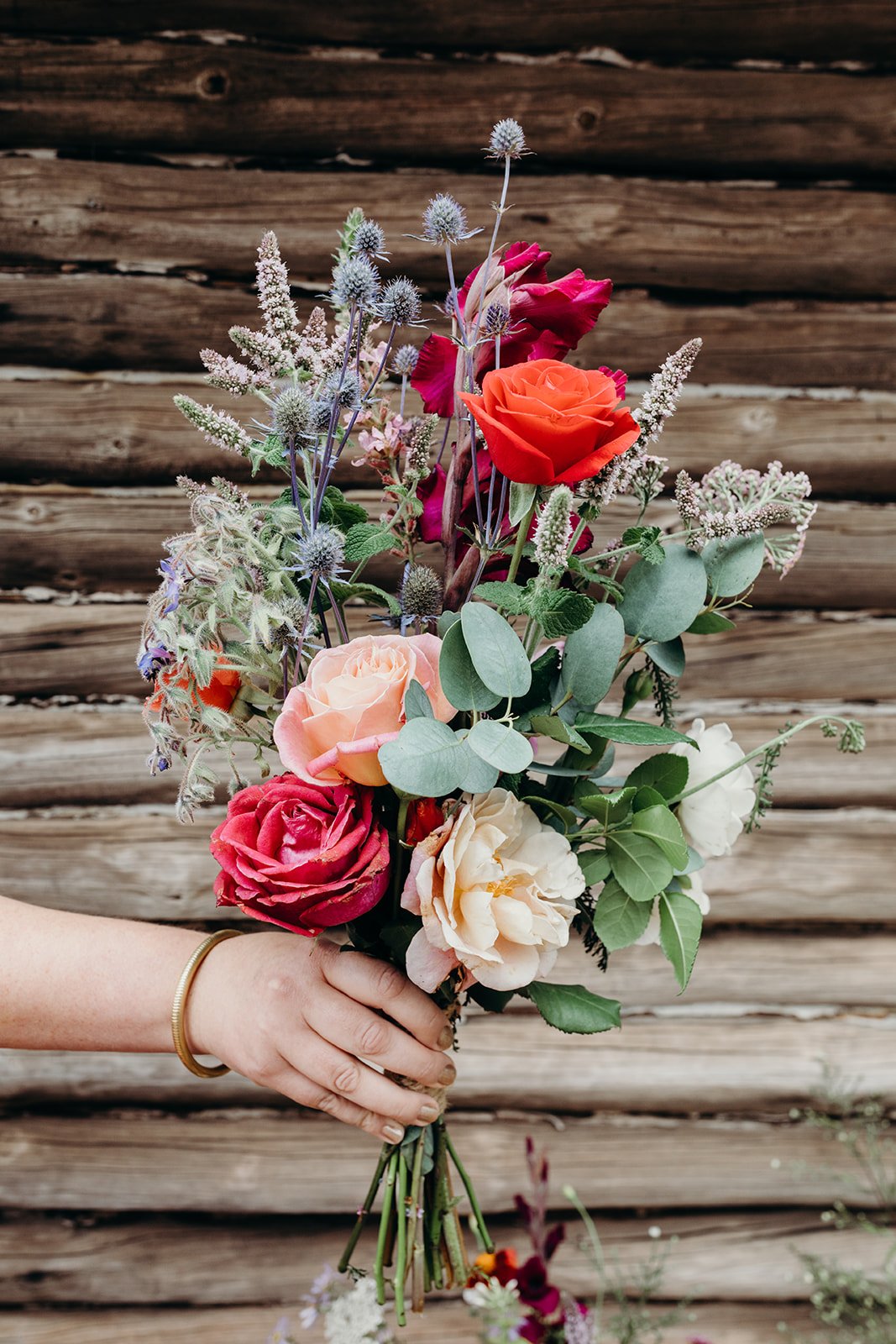 A person holding a colorful bouquet of roses and wildflowers in front of a log cabin wall.