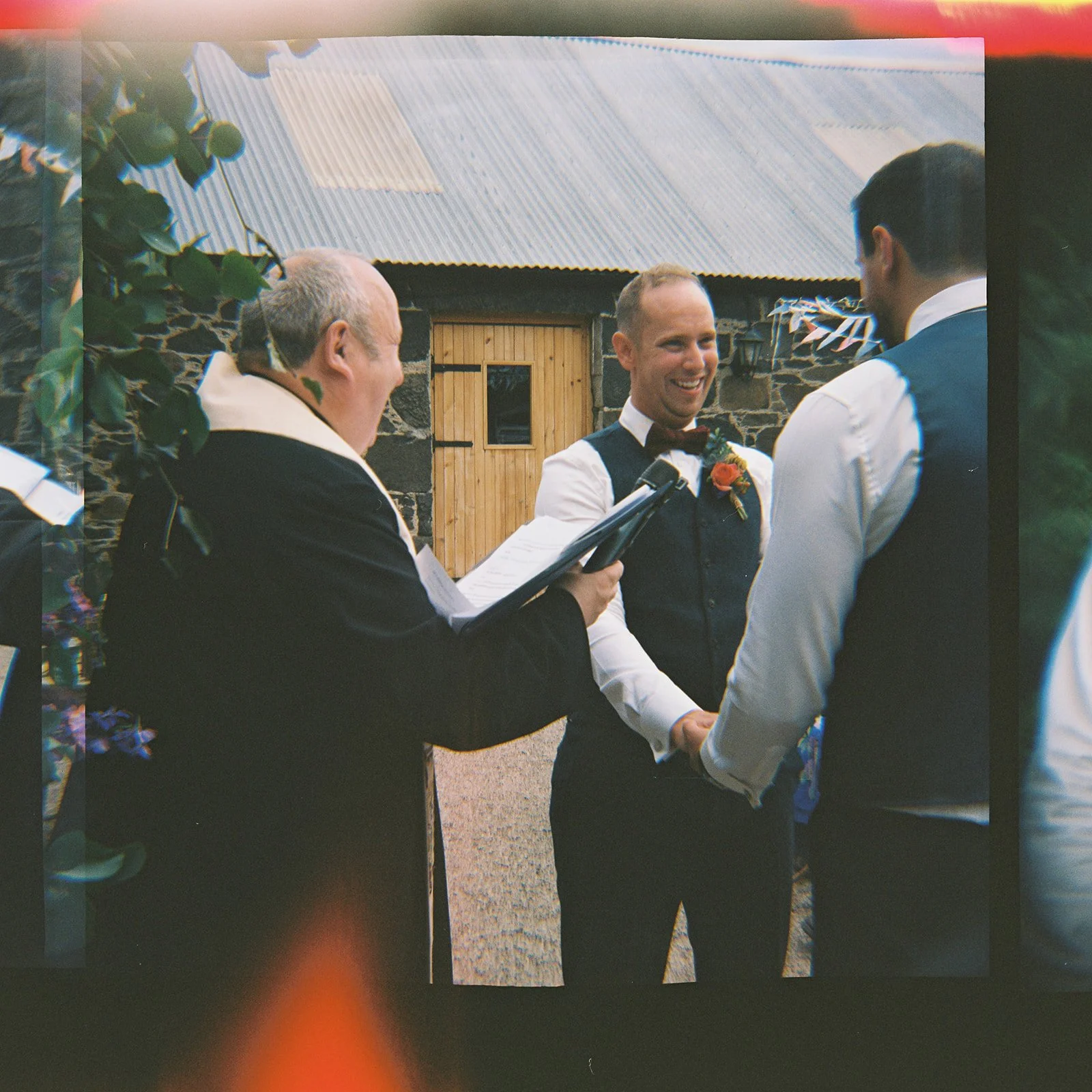 A groom and his partner holding hands during a wedding ceremony outdoors, with an officiant reading from a book. The groom is smiling, wearing a dark vest and bow tie, and the officiant is dressed in black, standing in front of a rustic stone buildin