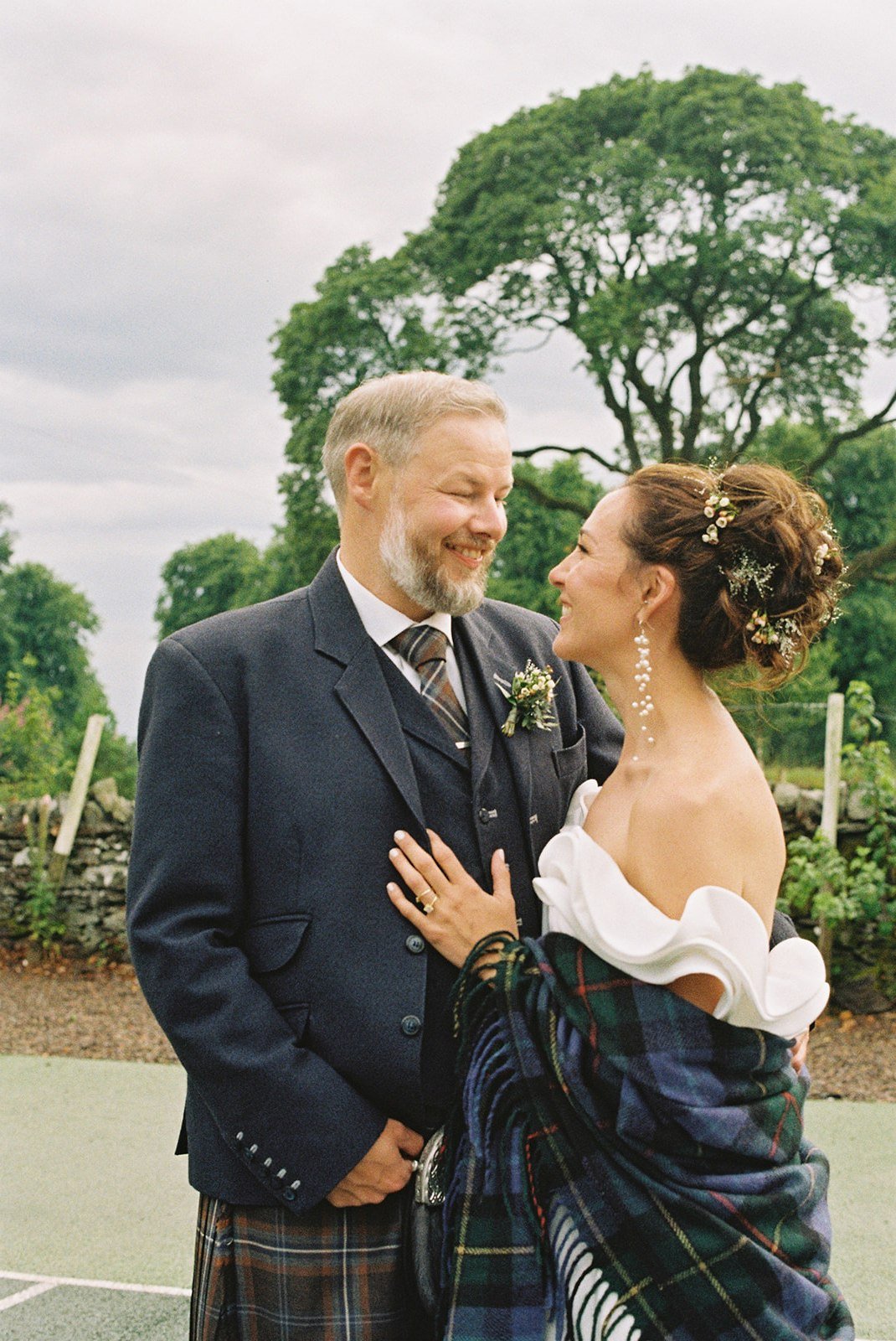 A bride and groom on their wedding day outdoors, smiling and gazing at each other, with greenery and trees in the background.