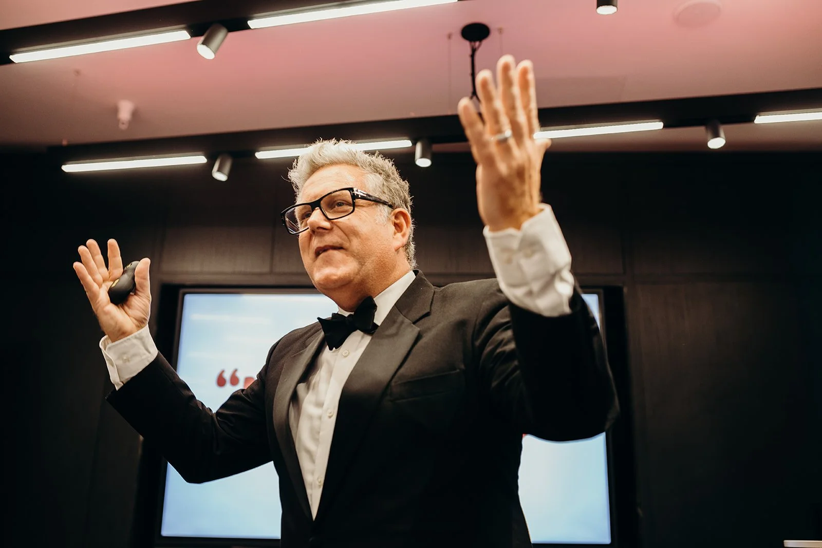 A man in a tuxedo and glasses, standing and gesturing with both hands, delivering a presentation or speech in a professional setting.