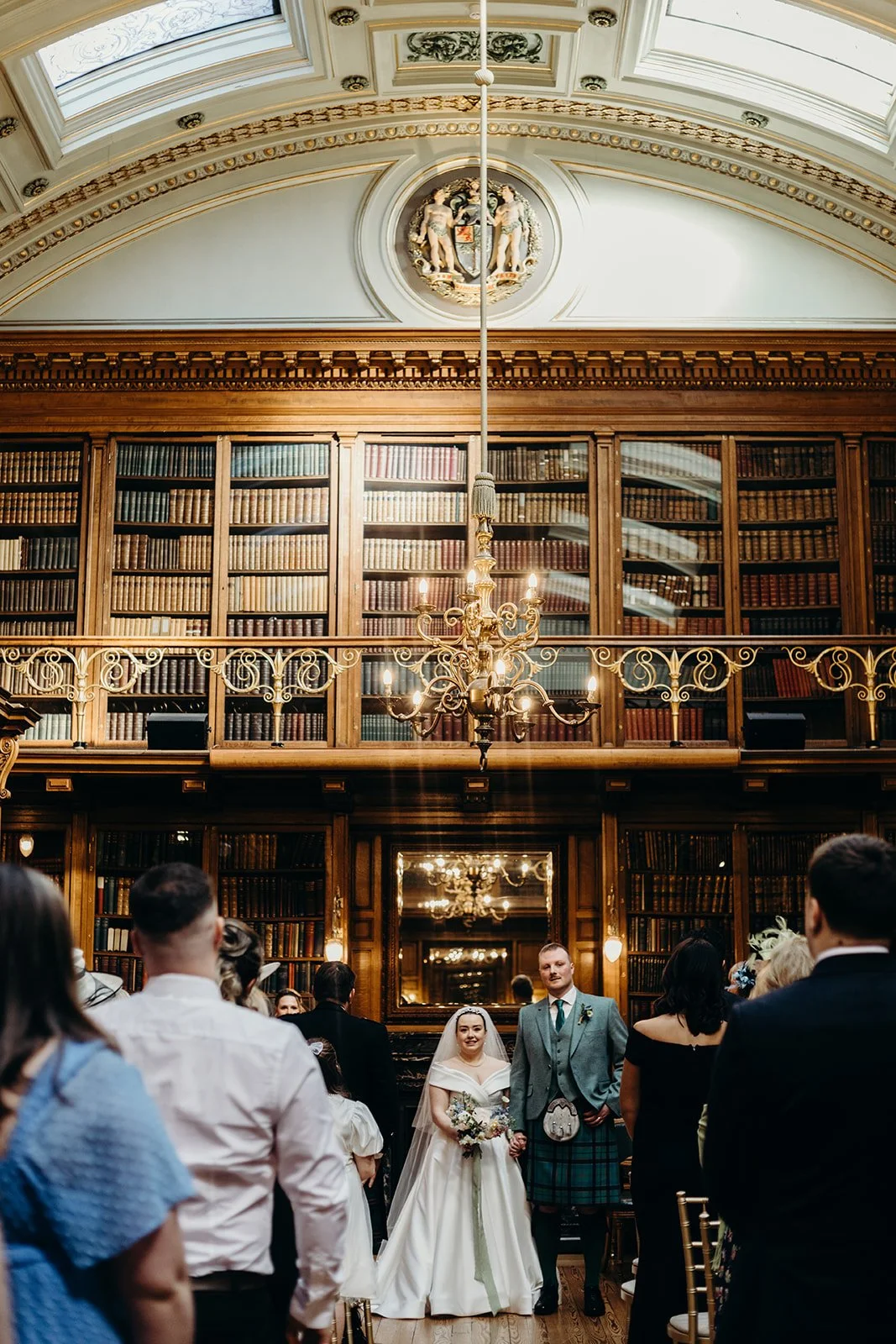 A wedding ceremony with a bride and groom standing hand in hand in a grand library room, surrounded by seated guests. The room features tall bookshelves, ornate chandeliers, and gold accents with a decorative ceiling and a mirror behind the couple.