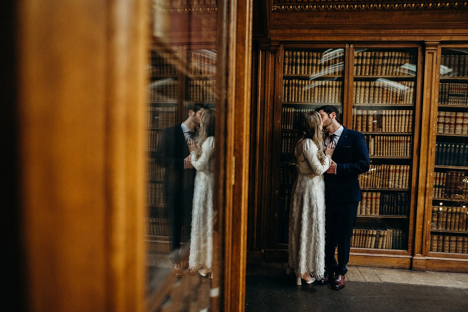 A couple dressed in formal attire sharing a kiss in a library filled with wooden bookshelves. The scene is reflected in a glass window, creating a mirrored effect.