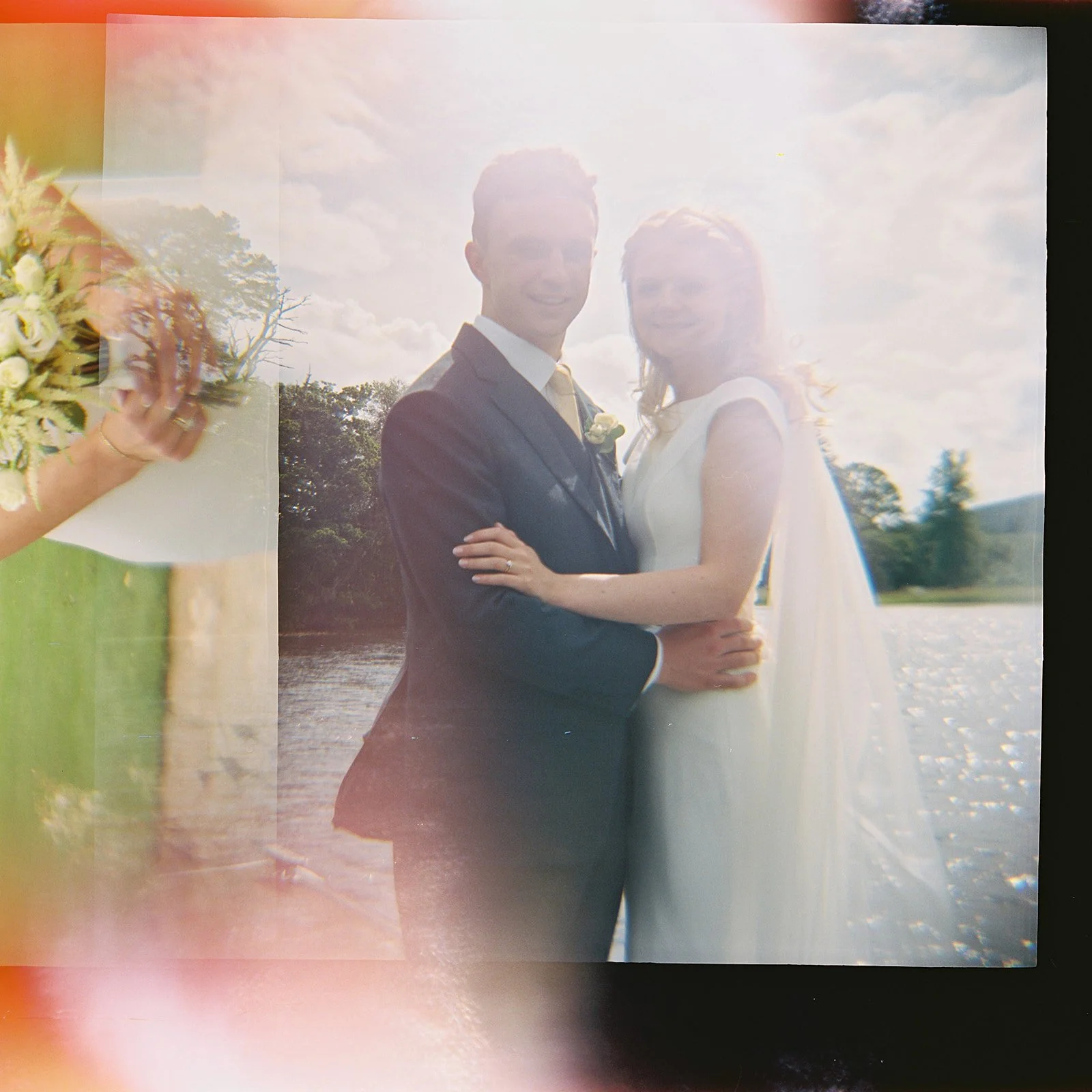 Photograph of a bride and groom standing together by a body of water outdoors on their wedding day.