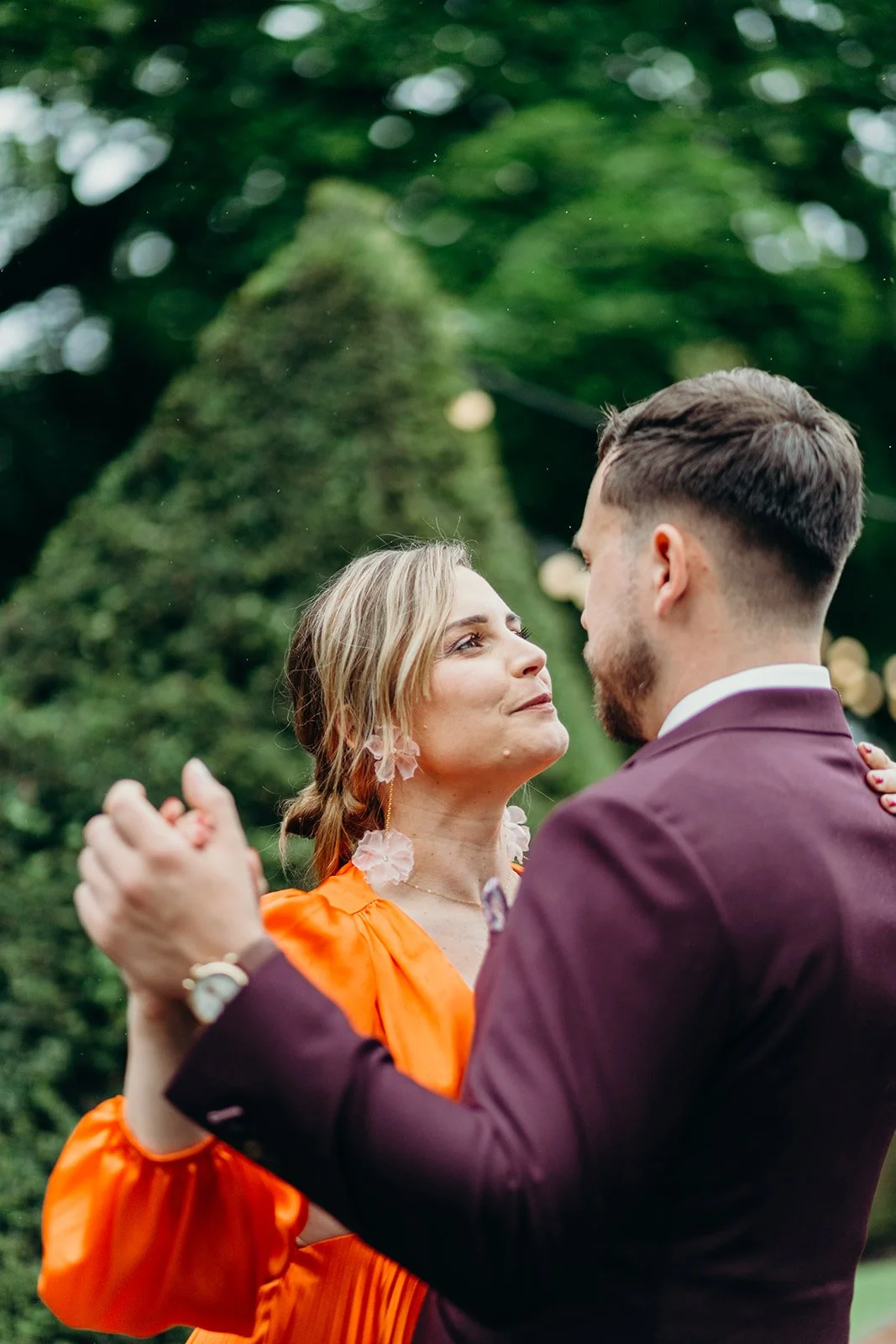 A woman and man dancing outdoors during sunset with greenery and a large bushy tree in the background.