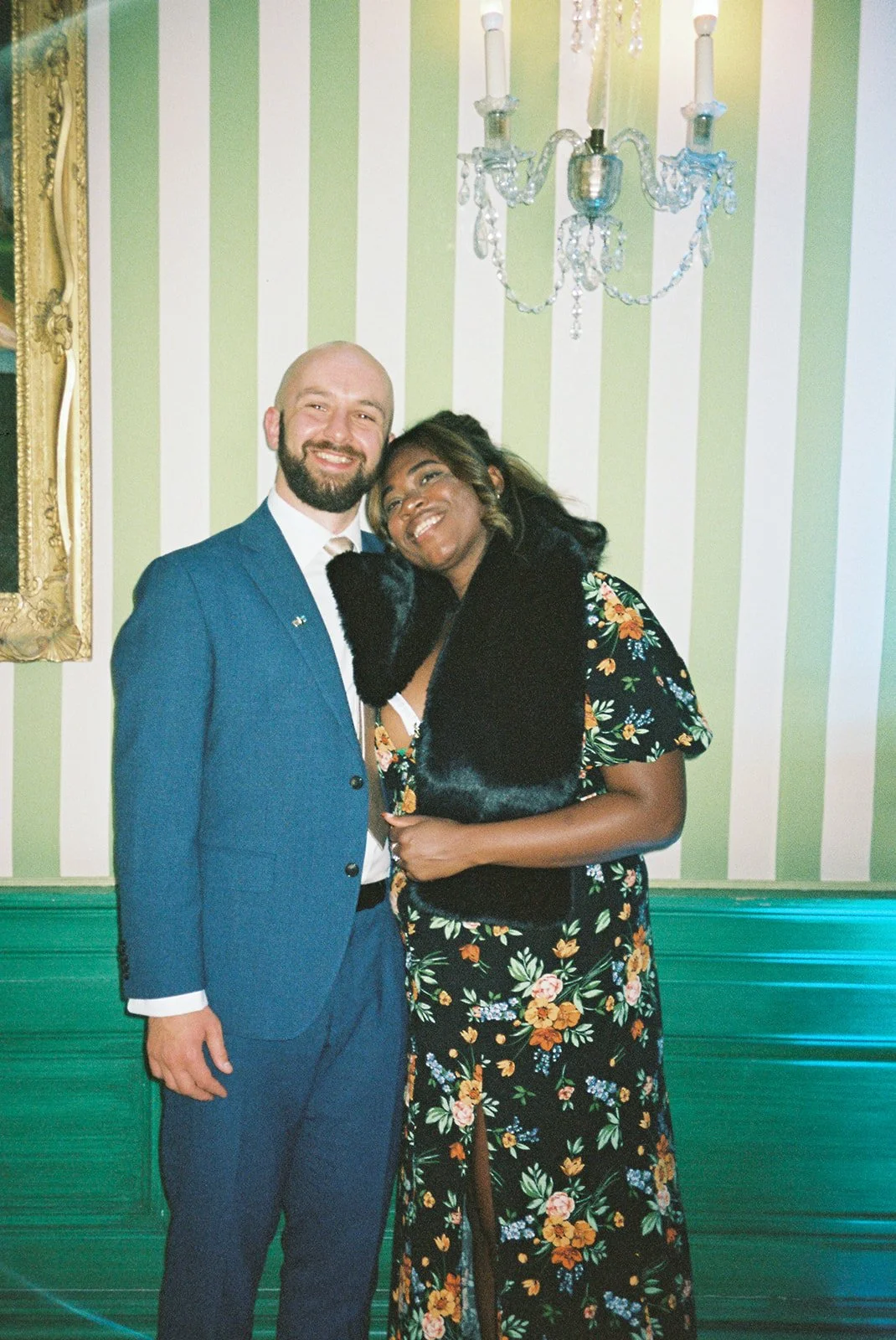 A smiling man and woman hugging, standing in a decorated room with green and white striped wallpaper and a chandelier overhead.