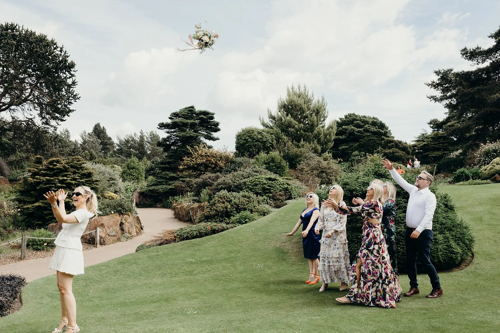 Group of people in a garden during a wedding or celebration, with a woman tossing a bouquet in the air.